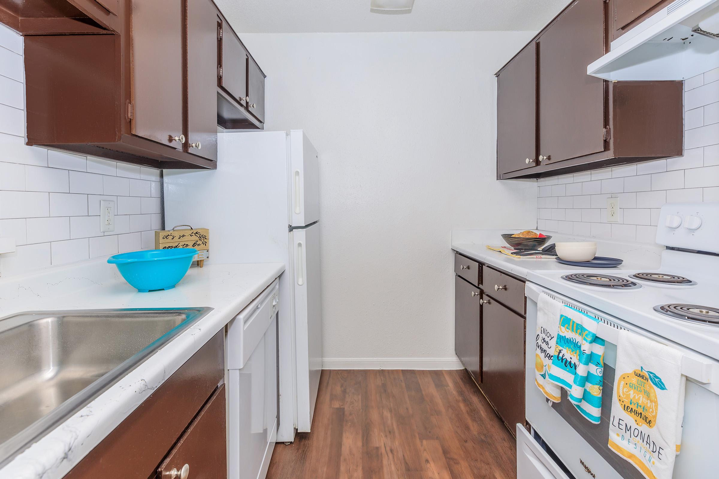 A clean, modern kitchen featuring dark wood cabinets, white countertops, and stainless steel appliances. There is a double sink on the left, a white refrigerator, and an oven on the right. A blue bowl sits on the counter, along with kitchen towels and a few cooking items, set against a backdrop of white tiled walls.