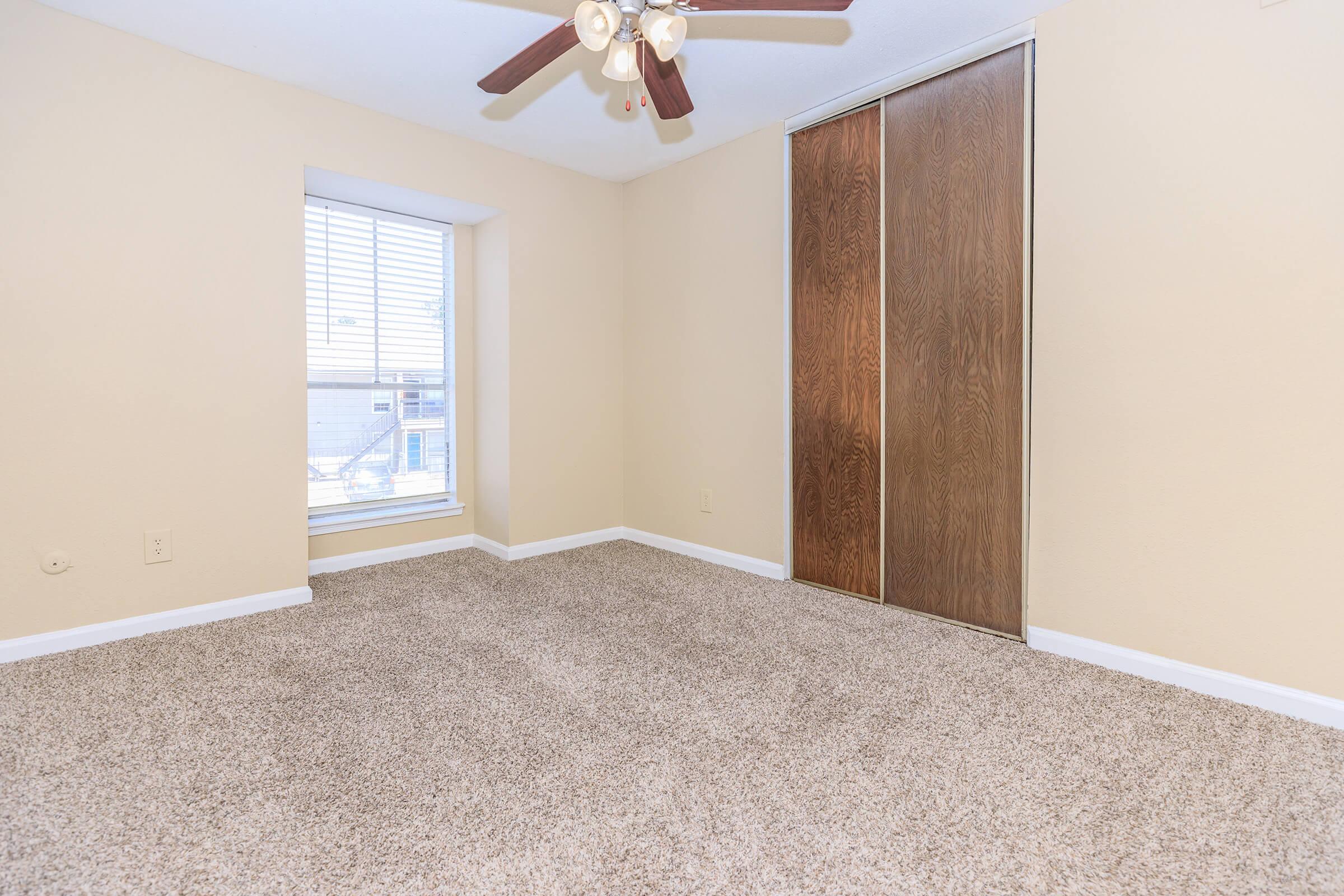 A cozy, empty bedroom featuring light beige walls and plush carpet. There is a window with white blinds letting in natural light, and a ceiling fan with wooden blades. A sliding closet door made of wood is on one wall, adding storage space to the room. The overall ambiance is bright and inviting.