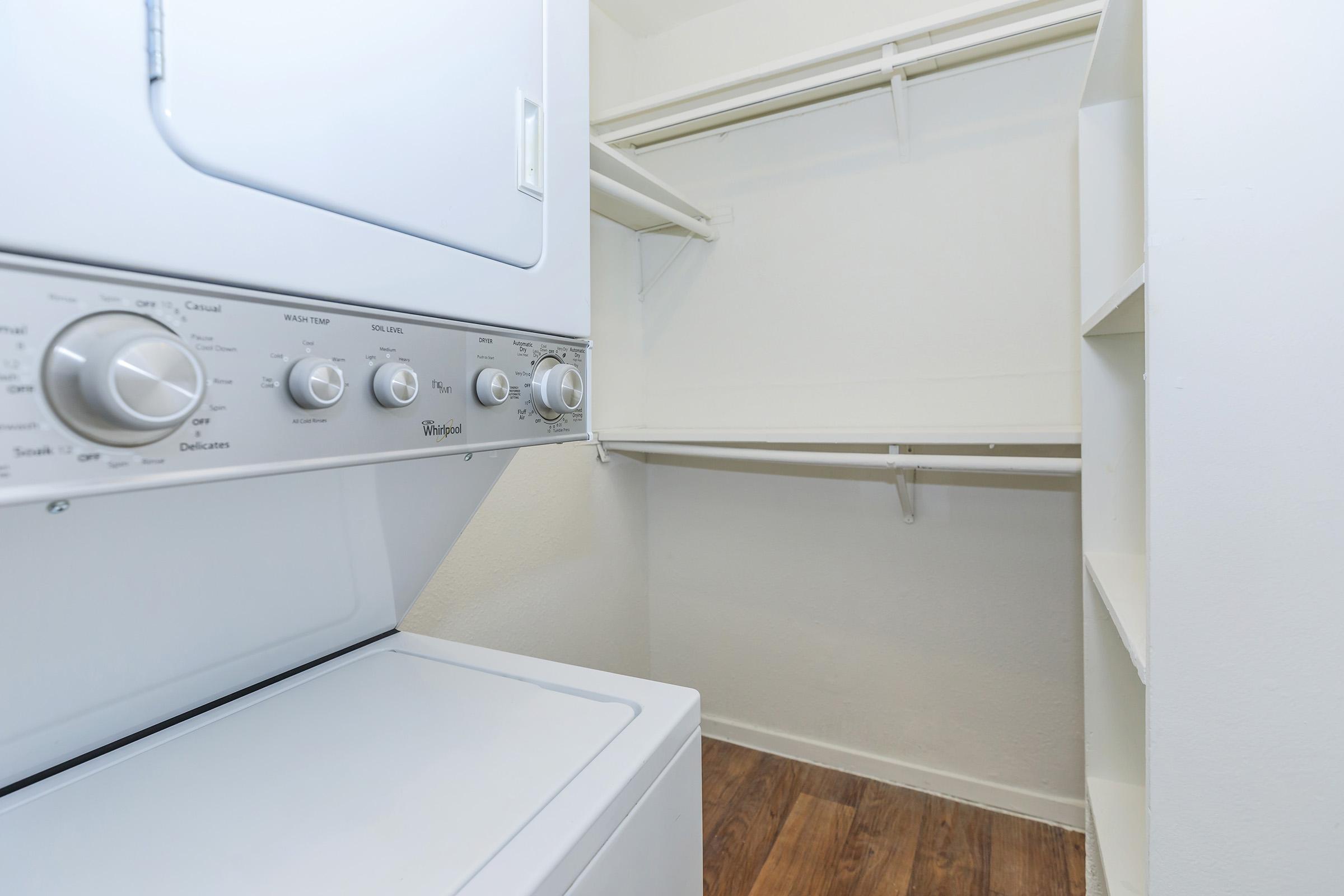 A clean laundry room featuring a stacked washer and dryer unit. The walls are painted light colors, and there are open shelves for storage. The floor has a wood-like finish, adding warmth to the space. A spacious closet area is visible, providing additional storage options.
