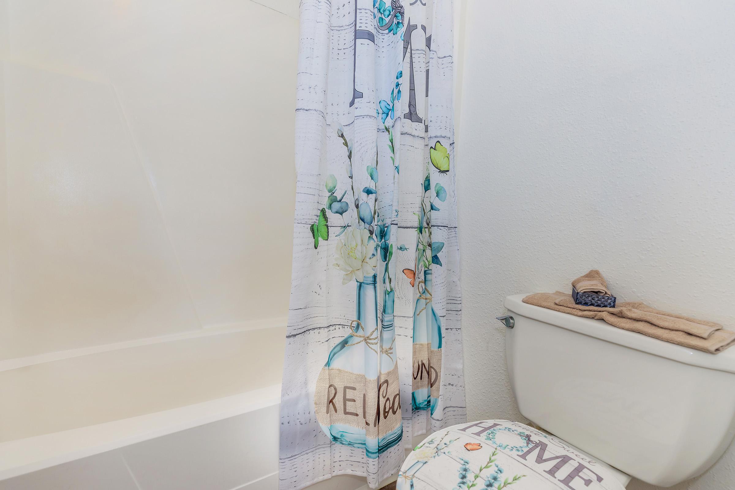 A bathroom featuring a shower with a decorative curtain displaying floral designs and bottles, a bathtub, and a toilet with a towel and decorative mat nearby. The walls are plain, and the overall decor is light and inviting.