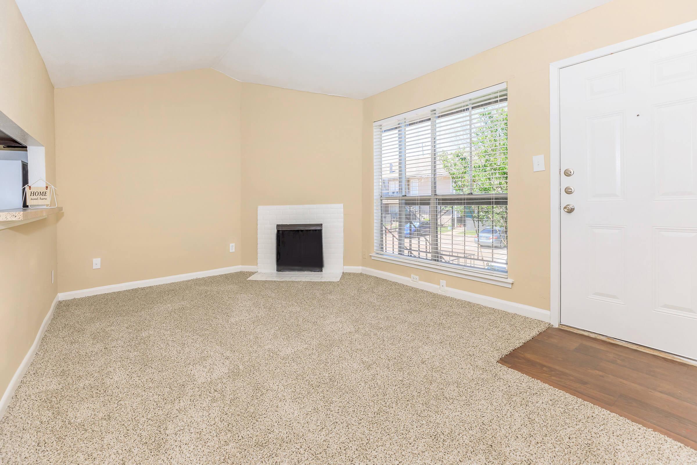 A spacious living room with light beige walls and carpet. It features a small white fireplace and large windows allowing natural light. There is a door on the right leading outside, and a cozy nook on the left, creating an inviting atmosphere.