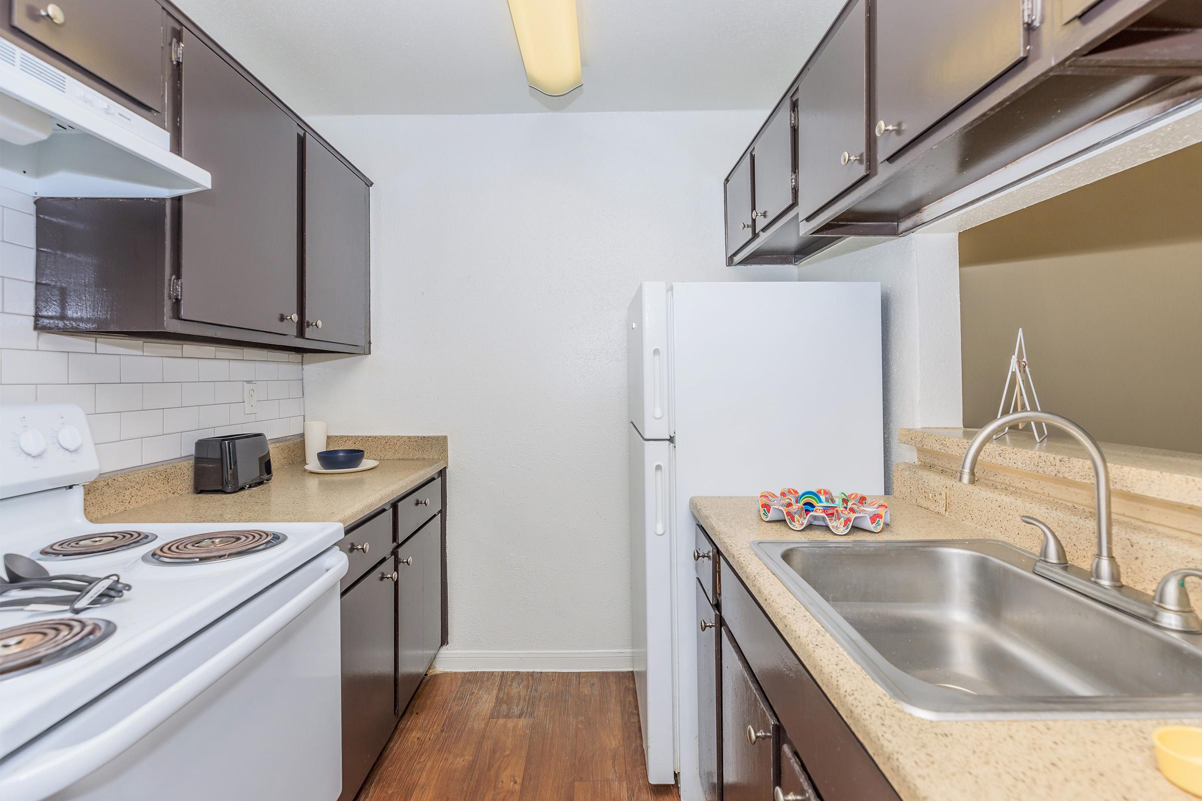A modern kitchen with dark cabinetry and a light-colored countertop. It features a white refrigerator, an electric stove with four burners, a microwave, and a double sink. The space is well-lit with a ceiling light, and there are some decorative items on the counter. The flooring is wood-like laminate.