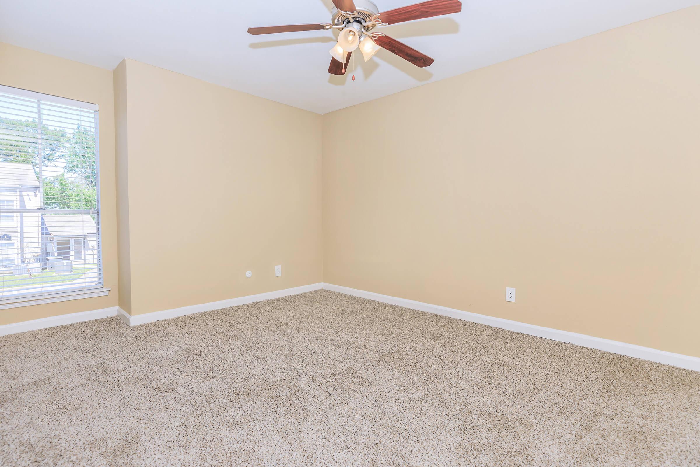 A spacious, empty room with beige walls and a light brown carpet. The room features a ceiling fan with wooden blades, and a window with blinds allows natural light to enter. There are no furnishings, creating an open and minimalistic feel.