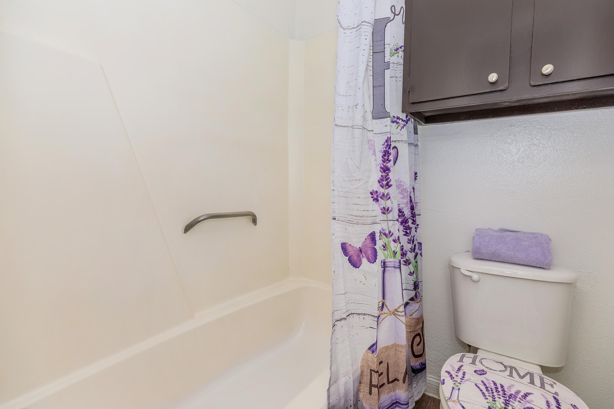 A bathroom with a shower area featuring a light-colored wall and a patterned shower curtain in purple and white. There is a toilet with a decorative cover that reads "HOME" and a folded purple towel on the tank. The overall decor gives a clean and homey feel.