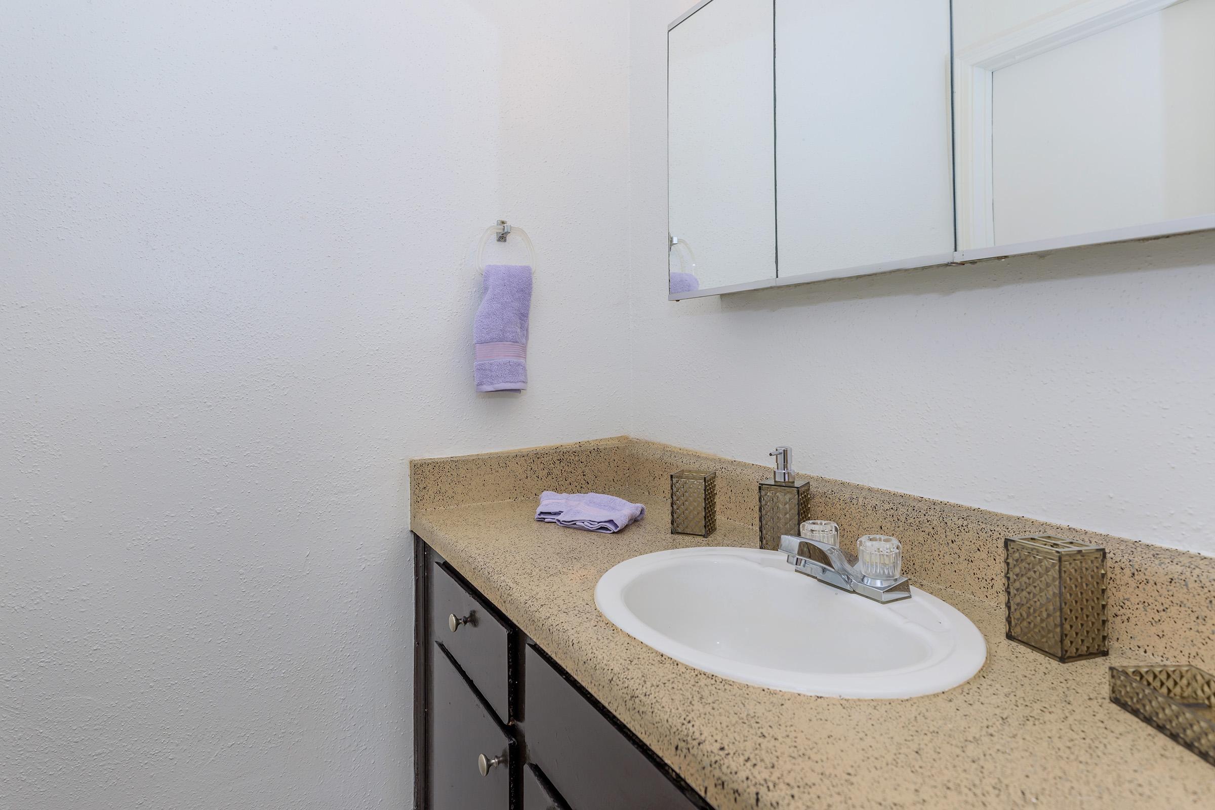 A clean bathroom scene featuring a beige countertop with a circular sink. There are two small decorative containers and a purple towel hanging on the wall. A large mirror is mounted above the sink, reflecting the simple, modern design. The walls are painted white, contributing to a fresh, airy atmosphere.