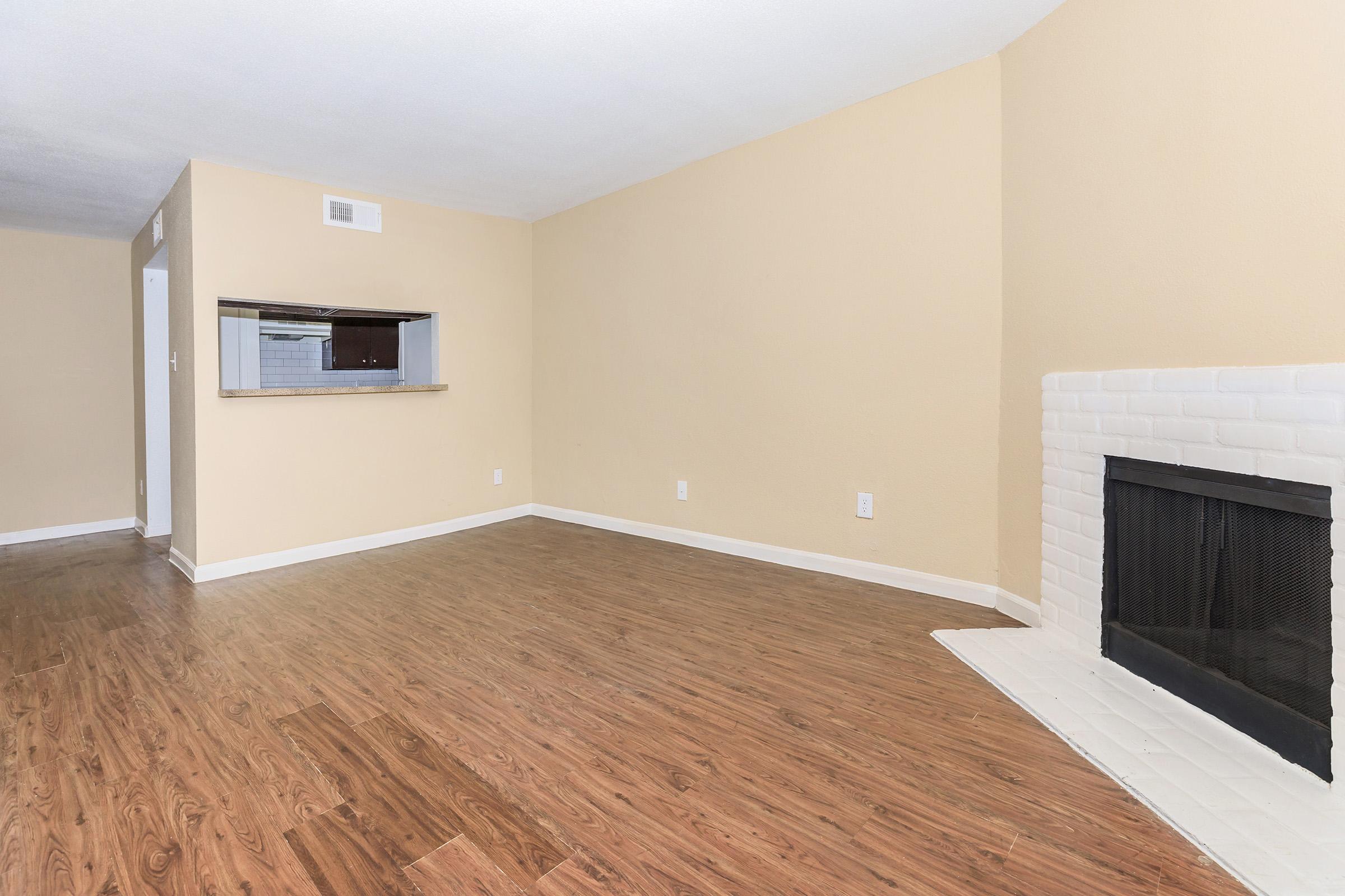 Spacious living room with light beige walls and laminate flooring. A white brick fireplace is on the right, and a partial view of an open kitchen is visible in the background. The room is well-lit, offering a cozy atmosphere with a neutral color palette.