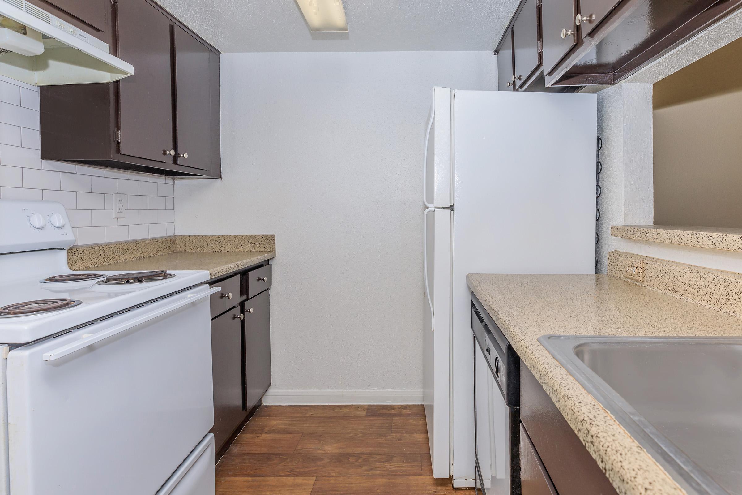 A modern kitchen featuring dark wood cabinets, a white stove and refrigerator, and a light-colored countertop. The walls are painted white, and there's a sink with a single faucet. The floor is made of brown wood-like material, creating a clean and functional cooking space.