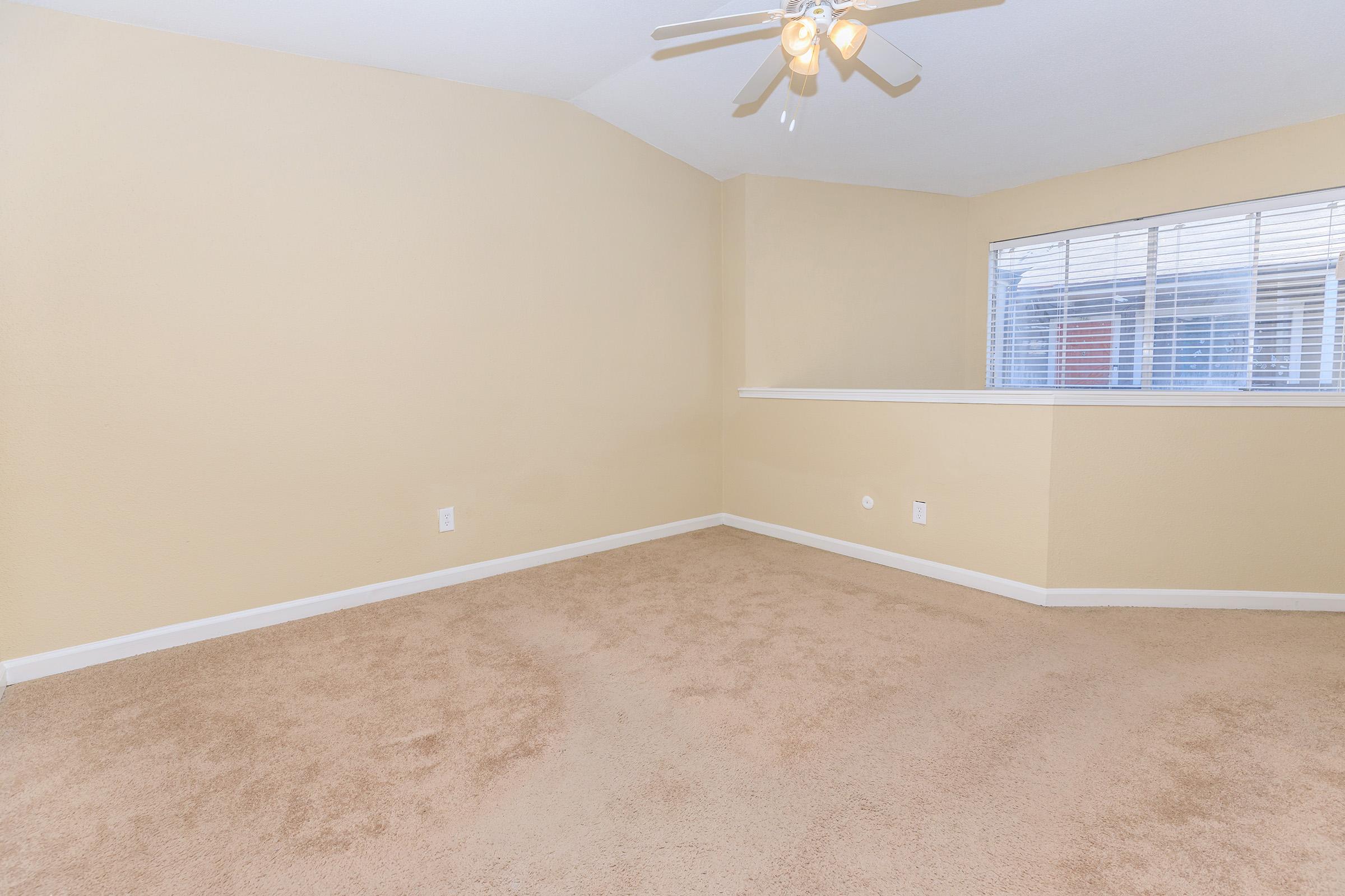 A spacious, empty room with light beige walls and carpeting. A ceiling fan is visible, and there is a large window with blinds allowing natural light to enter the space. The room features a slightly elevated section near the window, creating a cozy, open atmosphere suitable for various uses.