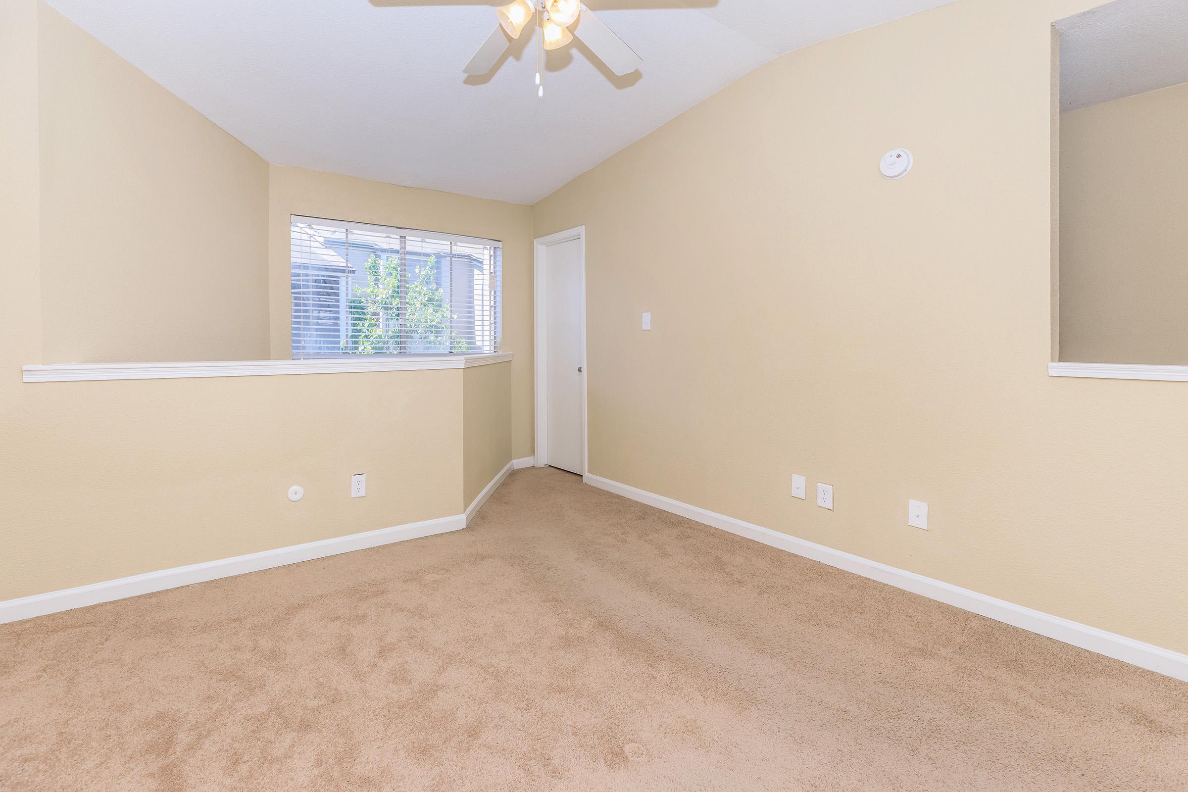 A spacious, empty room with beige walls and carpeted flooring. There is a ceiling fan, a window with natural light, and a door leading to another space. A small wall divides the room, creating an open but defined layout.