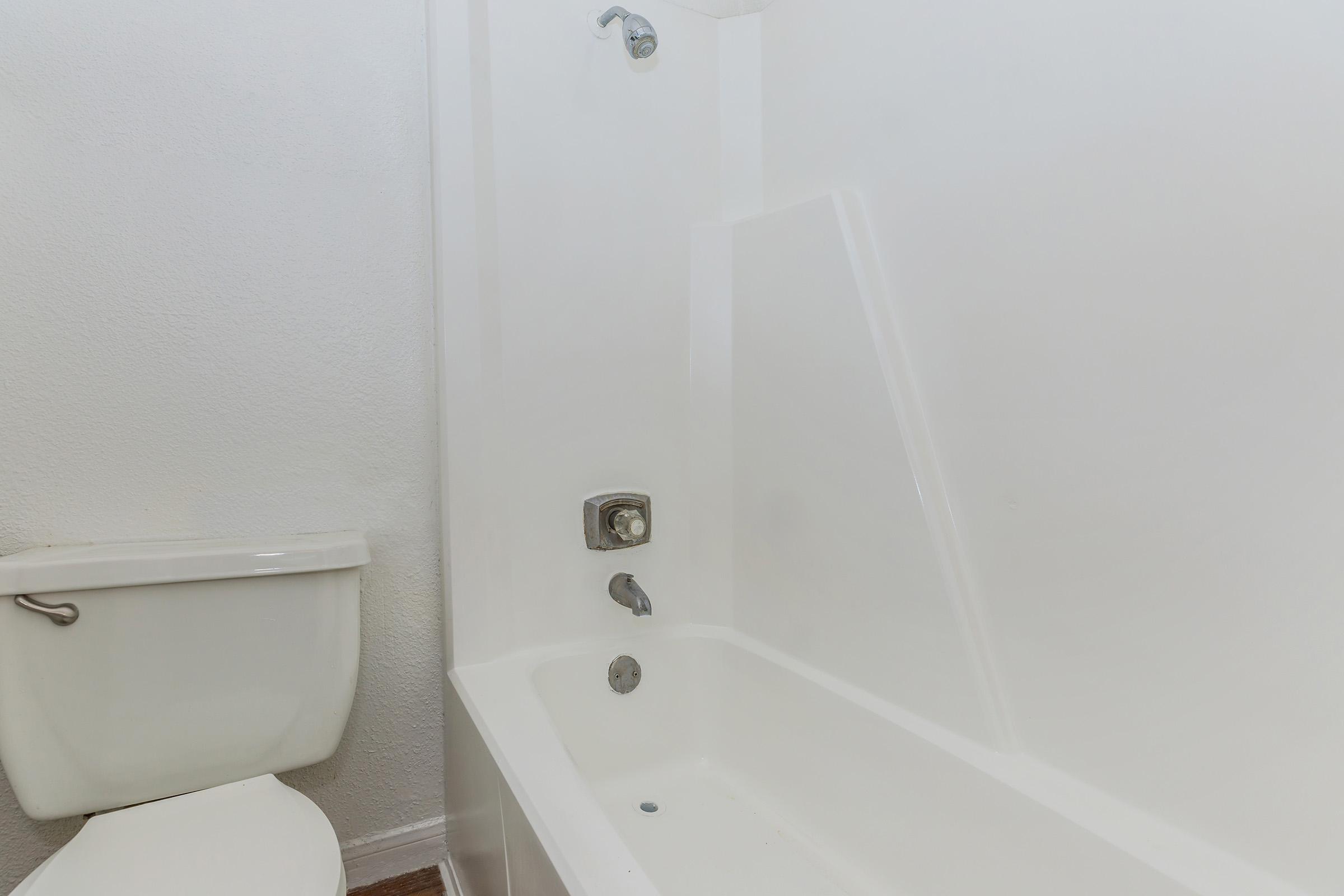 A clean and simple bathroom featuring a white bathtub with a showerhead, a faucet, and a separate white toilet nearby. The walls are light-colored, and the overall atmosphere is minimalistic and tidy.