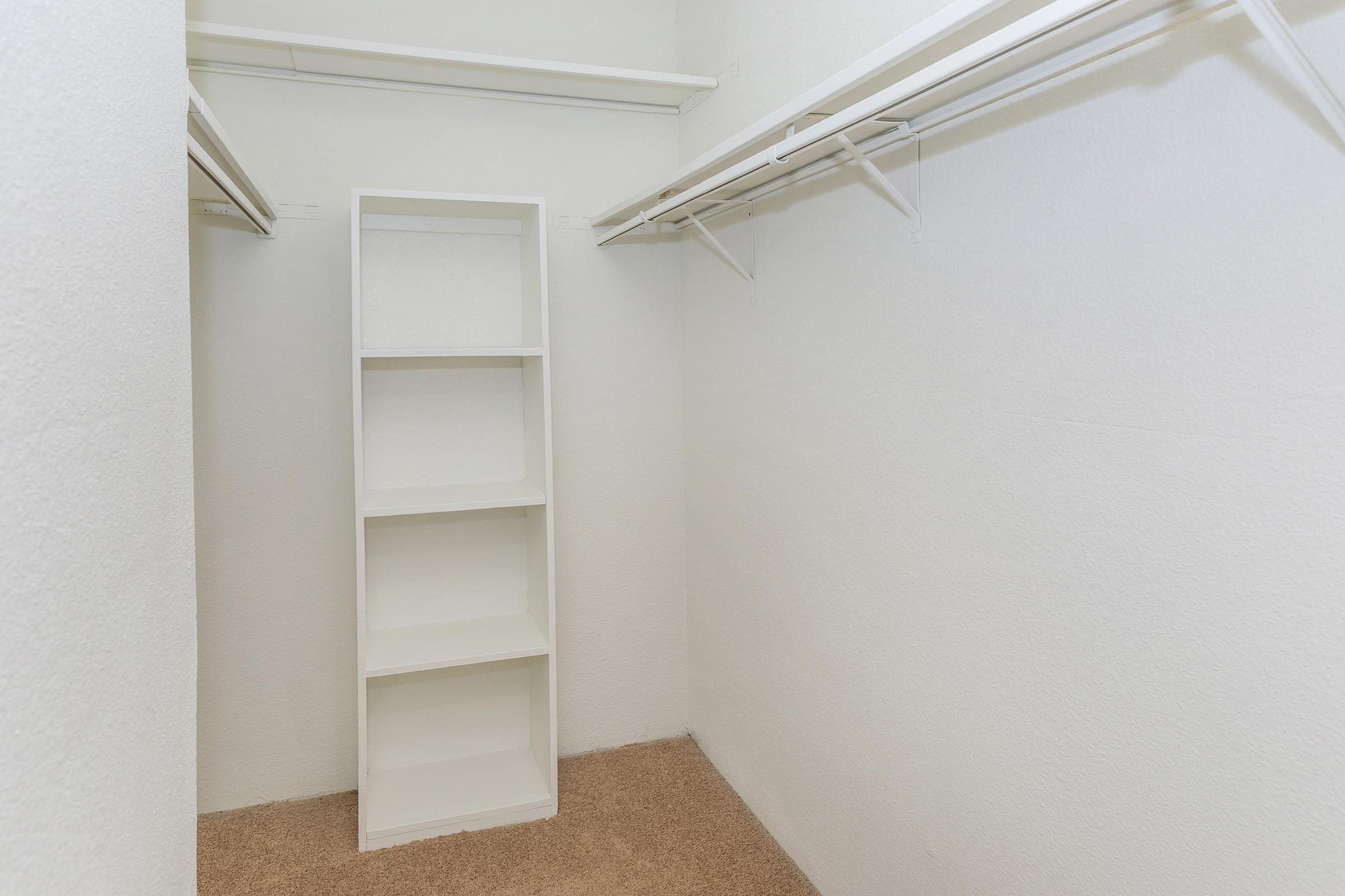 A clean, empty closet featuring white walls and a light brown carpeted floor. There is a tall, white shelving unit against one wall, with multiple open shelves, and two metal hanging rods for clothes on either side. The overall space is uncluttered and well-lit, giving a spacious appearance.