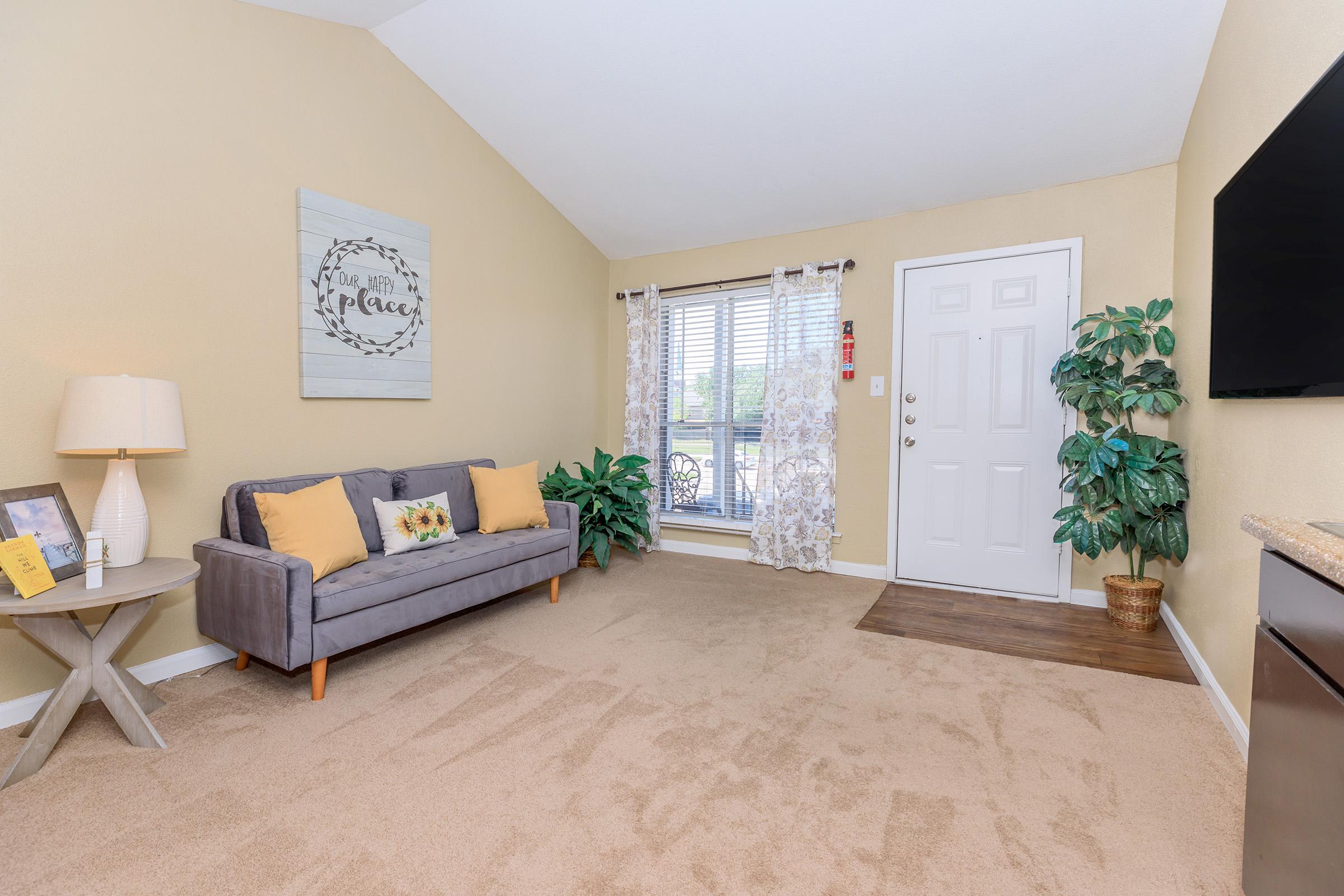 Cozy living room featuring a gray sofa with yellow cushions, a round side table, and a decorative lamp. There's a potted plant in the corner, a large window with sheer curtains, and a welcoming door. A wall-mounted TV is visible, along with a wall art piece that reads "Home is the best place to be."