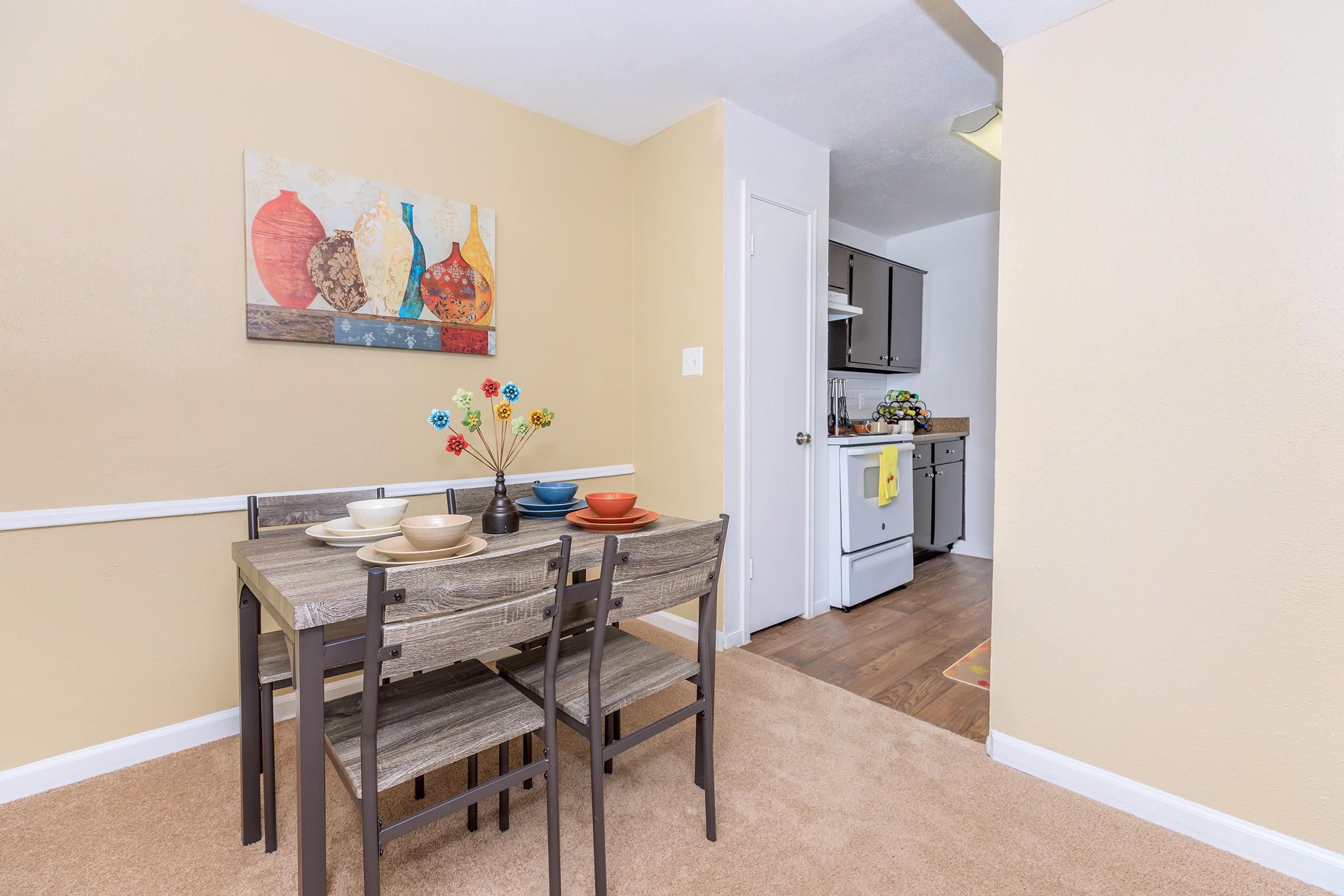 A cozy dining area featuring a small wooden table set with bowls in various colors. The wall is adorned with a vibrant painting of decorative vases. A door leads to a kitchen area, visible in the background, with dark cabinets and a colorful rug on the floor. The walls are painted a warm beige, enhancing the inviting atmosphere.