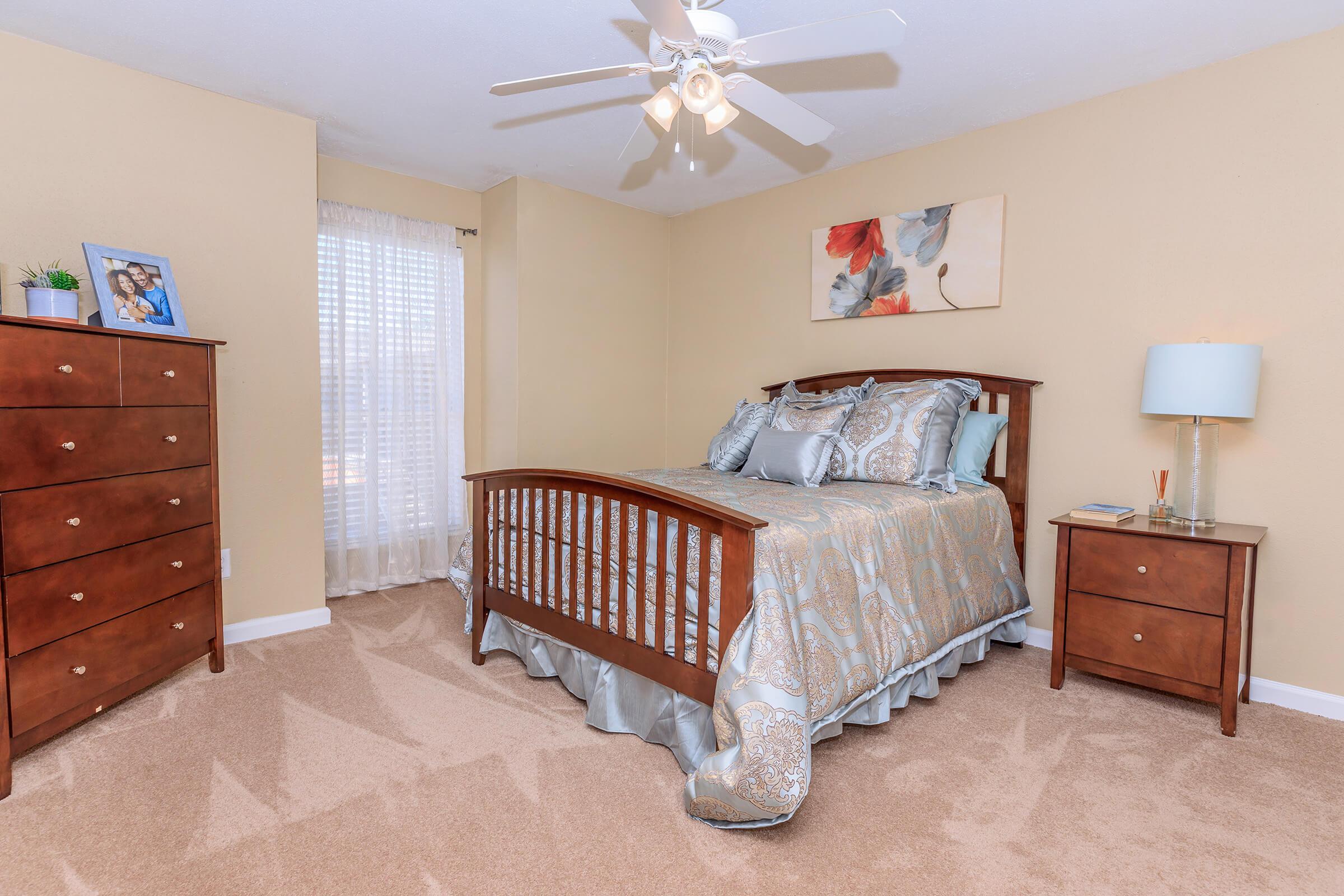 A cozy bedroom featuring a wooden bed with decorative bedding, a nightstand with a lamp, a dresser, and a framed photo on top. The walls are painted a warm beige, and natural light comes through a sheer curtain. A ceiling fan adds to the room's comfort.