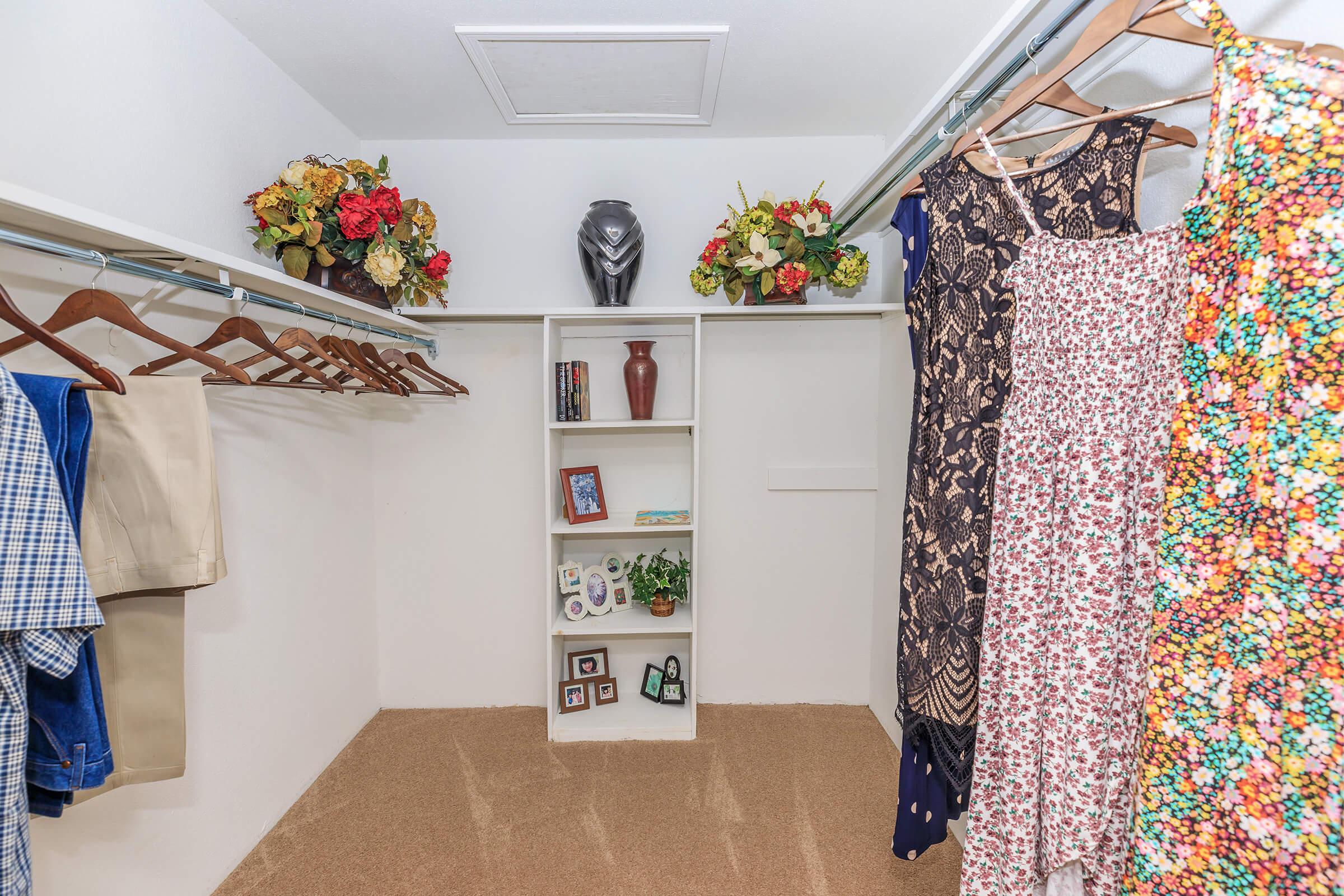 A spacious closet featuring hanging clothes on wooden hangers, including a floral dress and patterned garments. A central shelf displays decorative items, framed photos, and a vase, surrounded by faux flowers. The floor is carpeted, creating a cozy atmosphere.