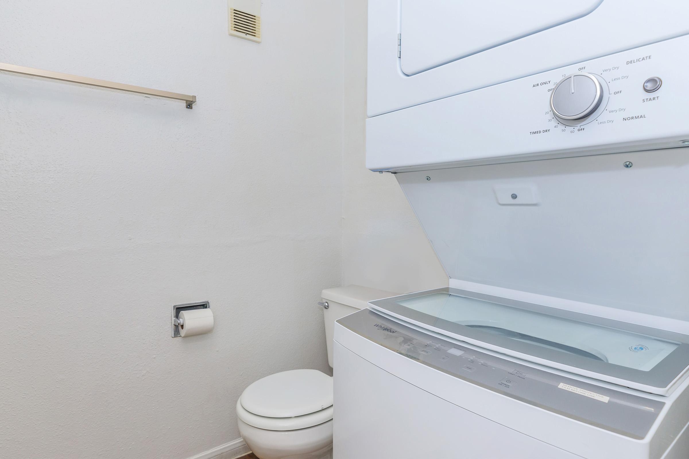 A compact laundry area featuring a stacked washer and dryer unit next to a white toilet. The walls are plain, and there is a towel rack mounted above the toilet. The floor is light-colored, enhancing the bright, clean appearance of the space.