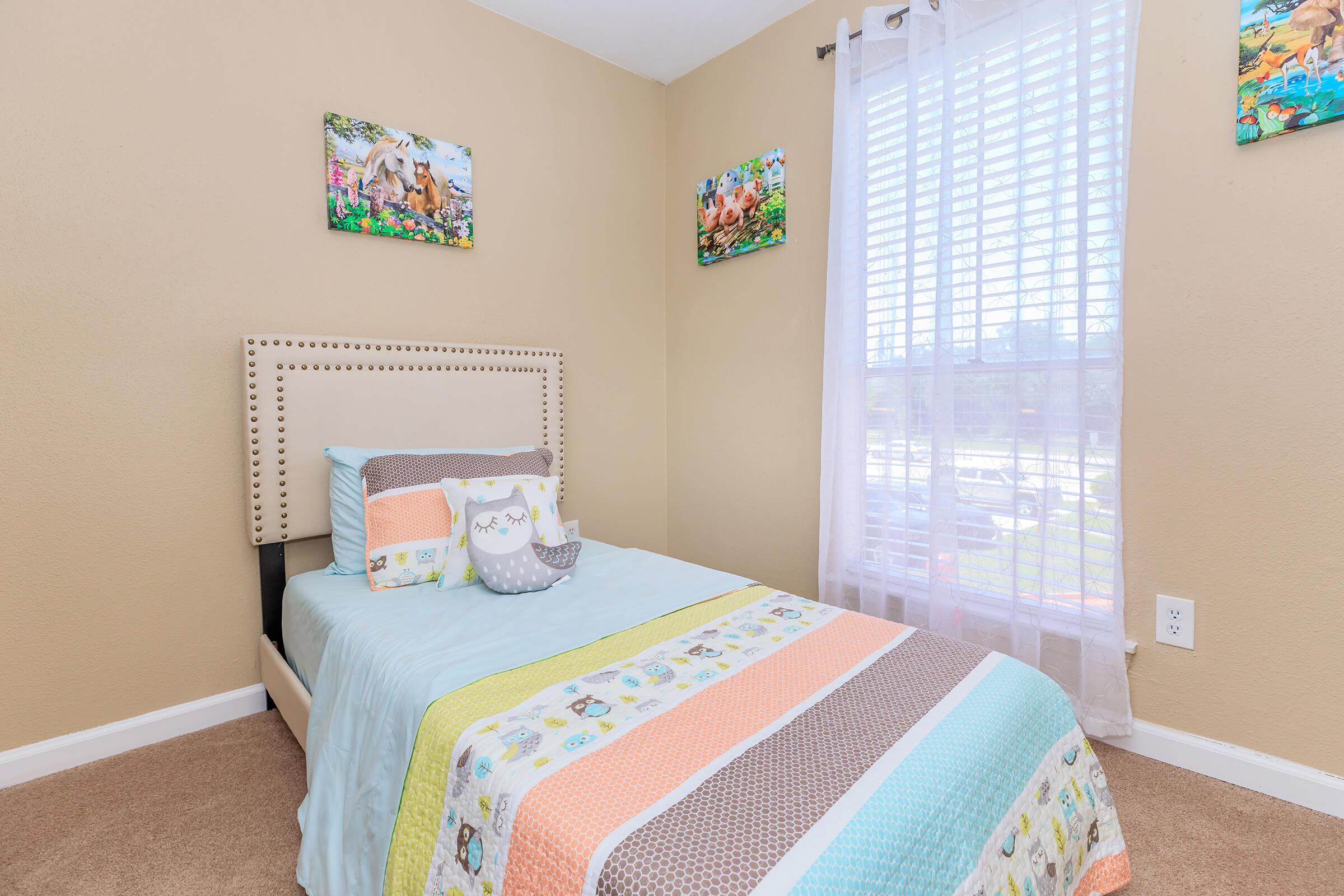 A cozy bedroom featuring a single bed with a colorful quilt, throw pillows, and light-colored walls. Sunlight streams through sheer curtains, illuminating the space. Two framed artwork pieces hang on the wall above the bed, adding a playful touch to the room. The carpet is beige, enhancing the inviting atmosphere.