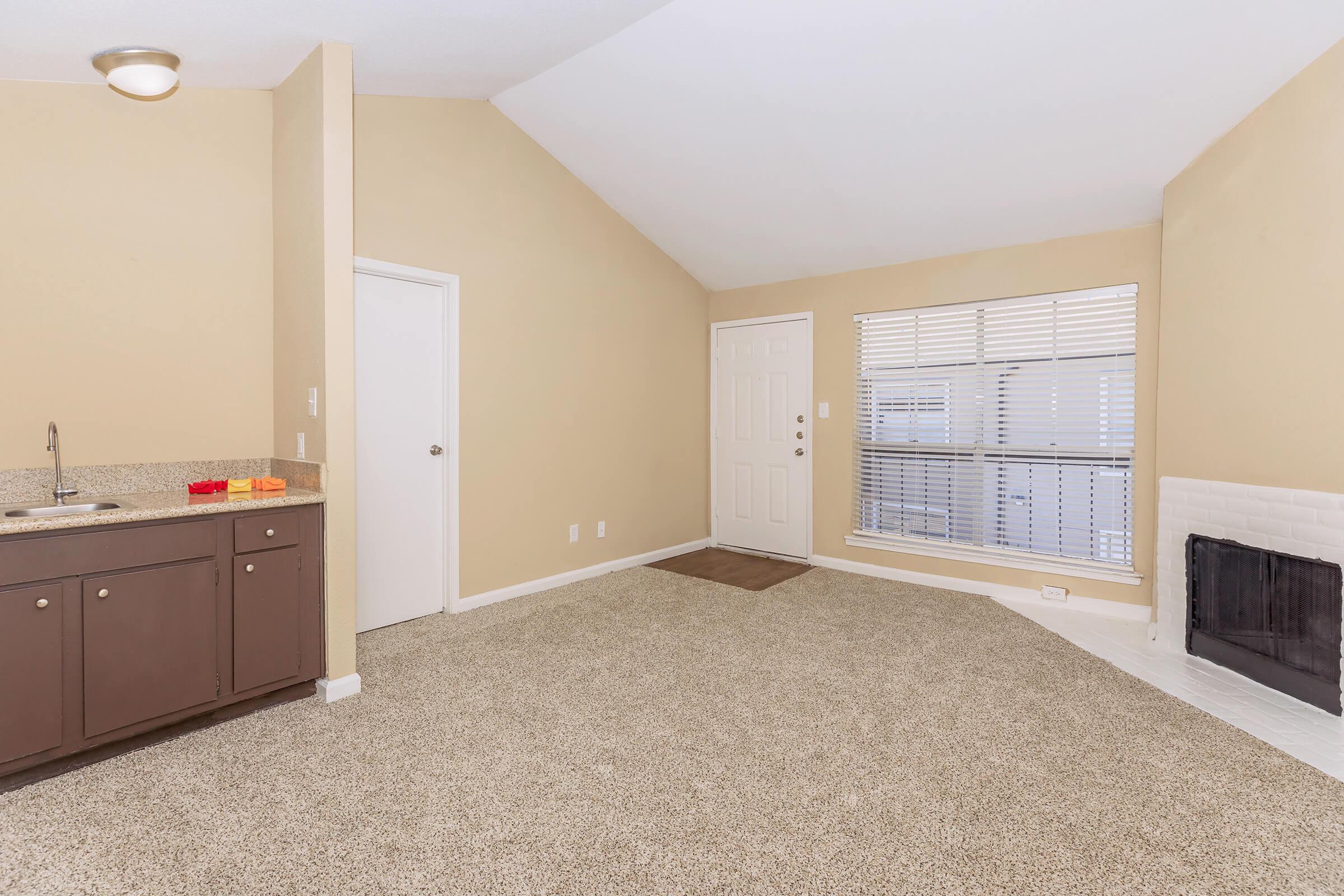 A spacious living area featuring light-colored walls, a vaulted ceiling, and a plush carpet. To the left is a kitchen area with dark cabinetry. A door leads outside, and large windows provide natural light. A modern fireplace is on the right side of the room. Overall, the space has a cozy and inviting atmosphere.