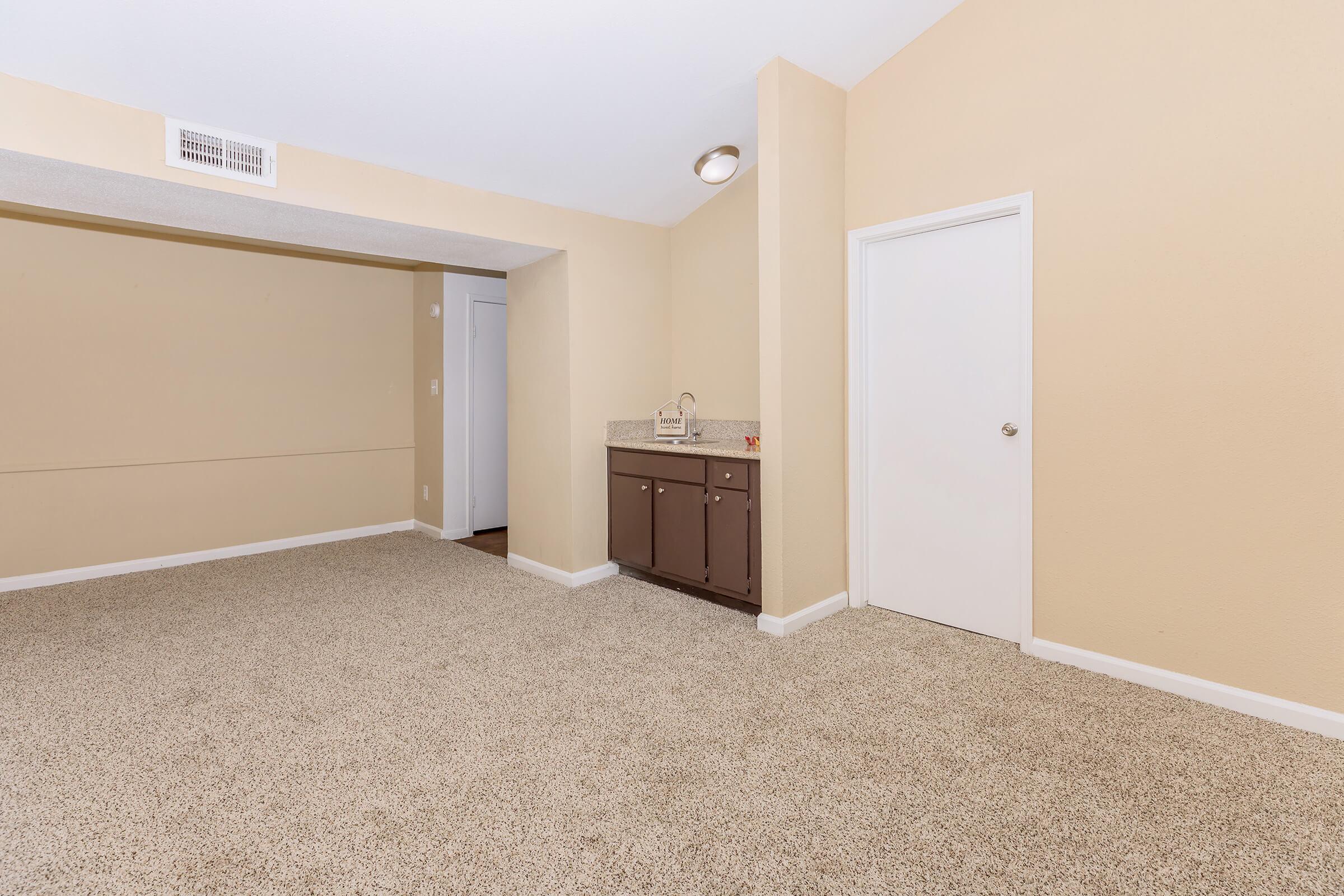 Empty room with beige walls, carpeted floor, and overhead lighting. There is a small built-in cabinet against one wall and a white door leading to another space. The overall atmosphere is bright and open, with no furniture or decorations present.