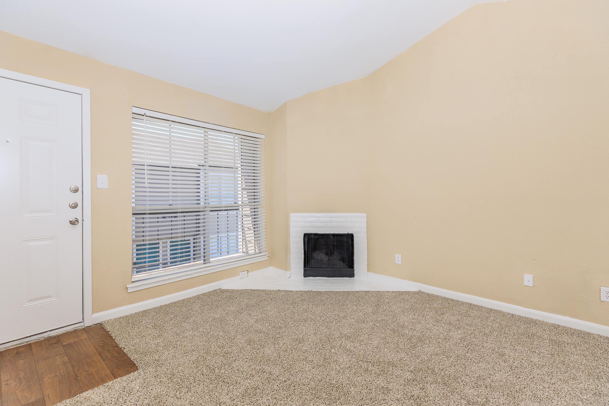 A cozy, empty living room with light beige walls and a large window featuring blinds. The floor is covered with soft, light brown carpet. In one corner, there is a white fireplace, and a front door is visible on the left side of the room. The space feels bright and inviting.