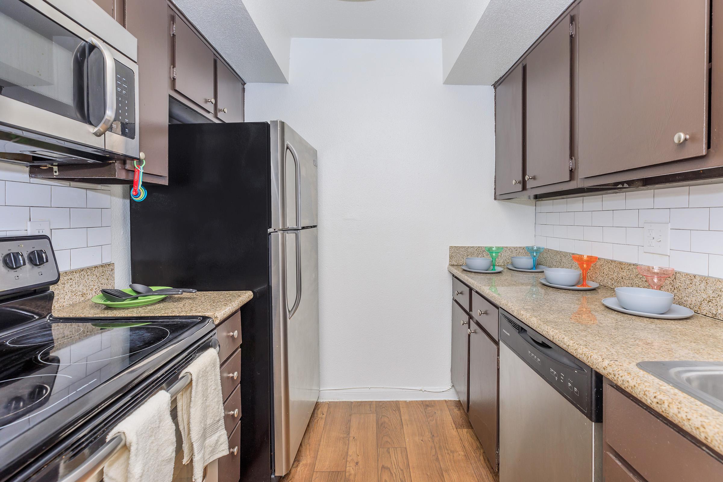 A modern kitchen featuring dark cabinetry, stainless steel appliances, and a granite countertop. The stove and microwave are visible, along with a silver refrigerator. Various colorful bowls are placed on the counter, and the floor has a warm wooden finish, creating a cozy atmosphere.