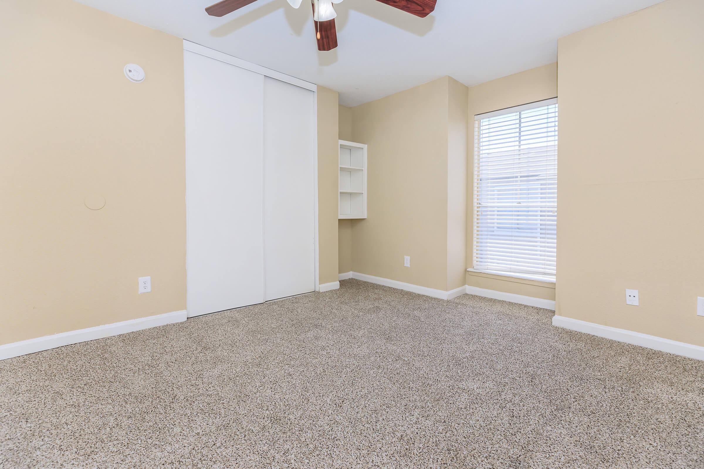 Spacious room featuring beige walls, carpet flooring, a ceiling fan, and a large window with blinds. Includes a built-in shelf and a closet with sliding doors. Natural light enhances the inviting atmosphere.