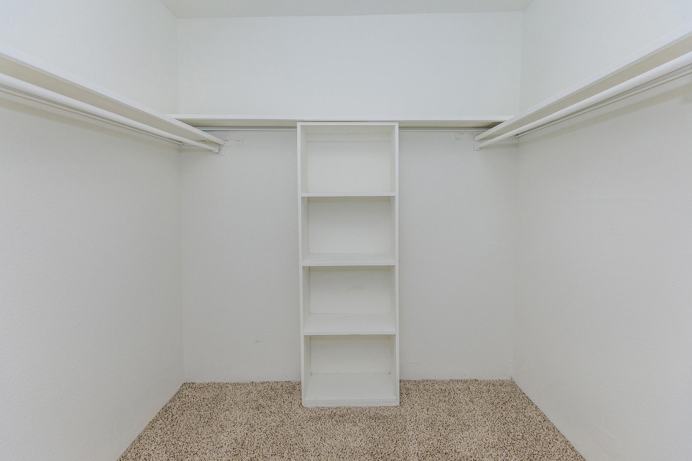 Empty closet space featuring plain white walls and a beige carpeted floor. There are two hanging rods on either side and a central white shelving unit with four open shelves. The overall appearance is clean and minimalistic, providing ample storage potential.