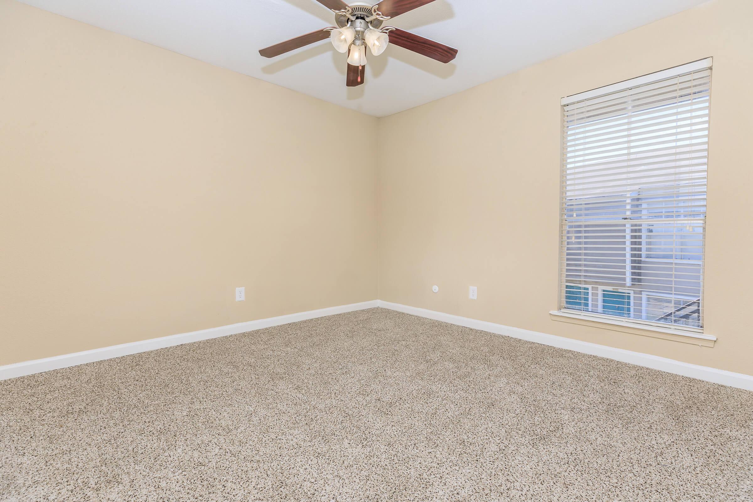 Empty room with beige walls and a light brown carpet. A ceiling fan with five blades is installed in the center, and there is a window with white blinds on one wall. The space is well-lit and uncluttered, offering a clean and neutral environment.