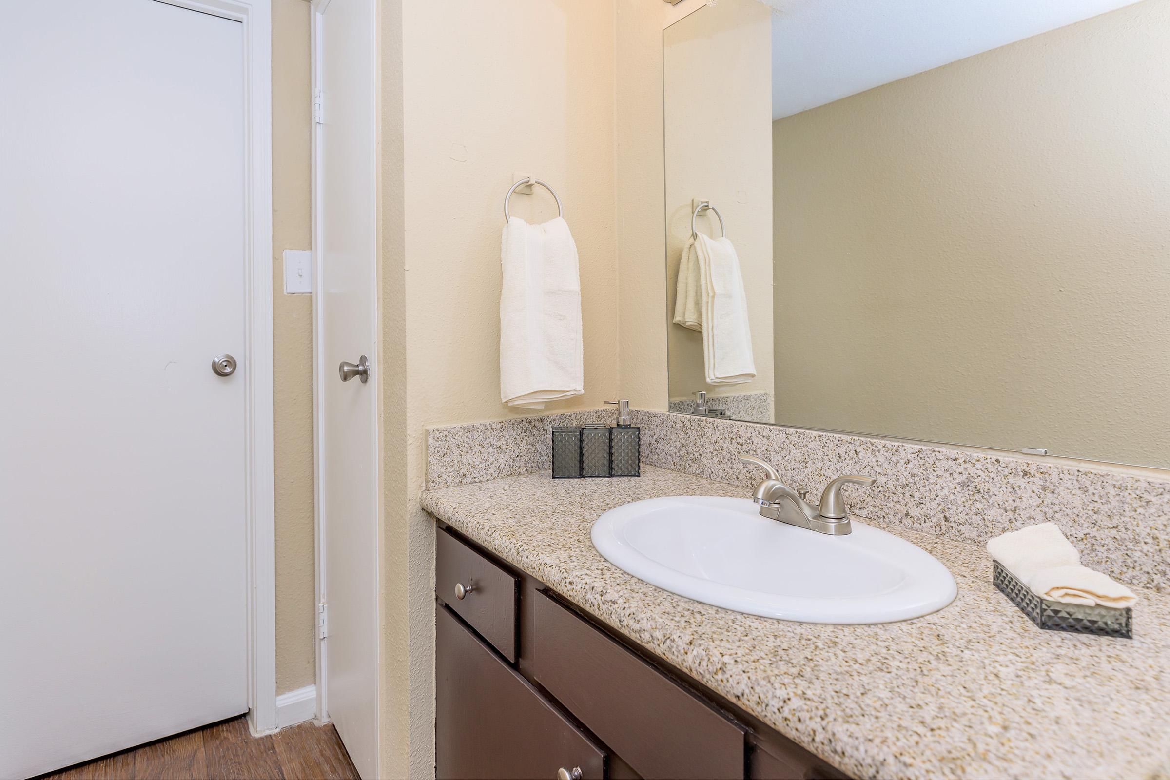 A clean bathroom with a light beige color scheme. The counter features a white sink, a soap dispenser, and a hand towel holder. A towel hangs on the wall, and a mirror is mounted above the sink. A closed door is visible on the left side of the image. The flooring is wooden.