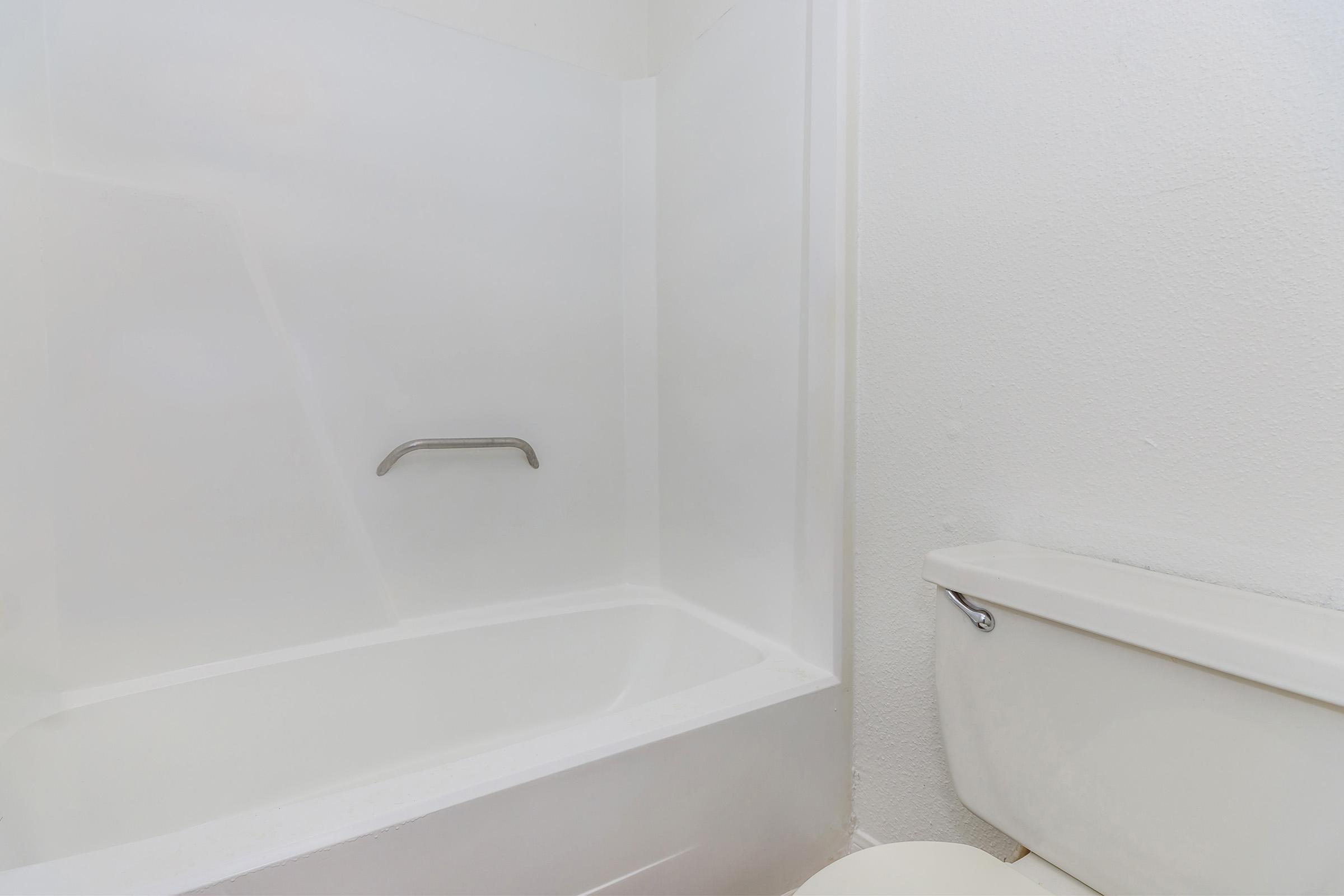 A clean, modern bathroom featuring a white bathtub with a grab bar and a white toilet nearby. The walls are light-colored, creating a bright and fresh atmosphere.