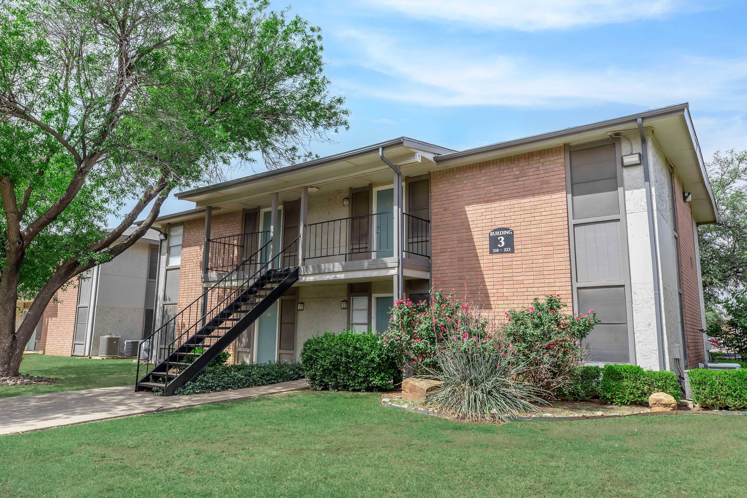Two-story apartment building with a brick facade and a black metal staircase leading to the upper level. The building is surrounded by grassy areas and a few trees. Flowering shrubs add color to the landscape. A clear blue sky is visible overhead.