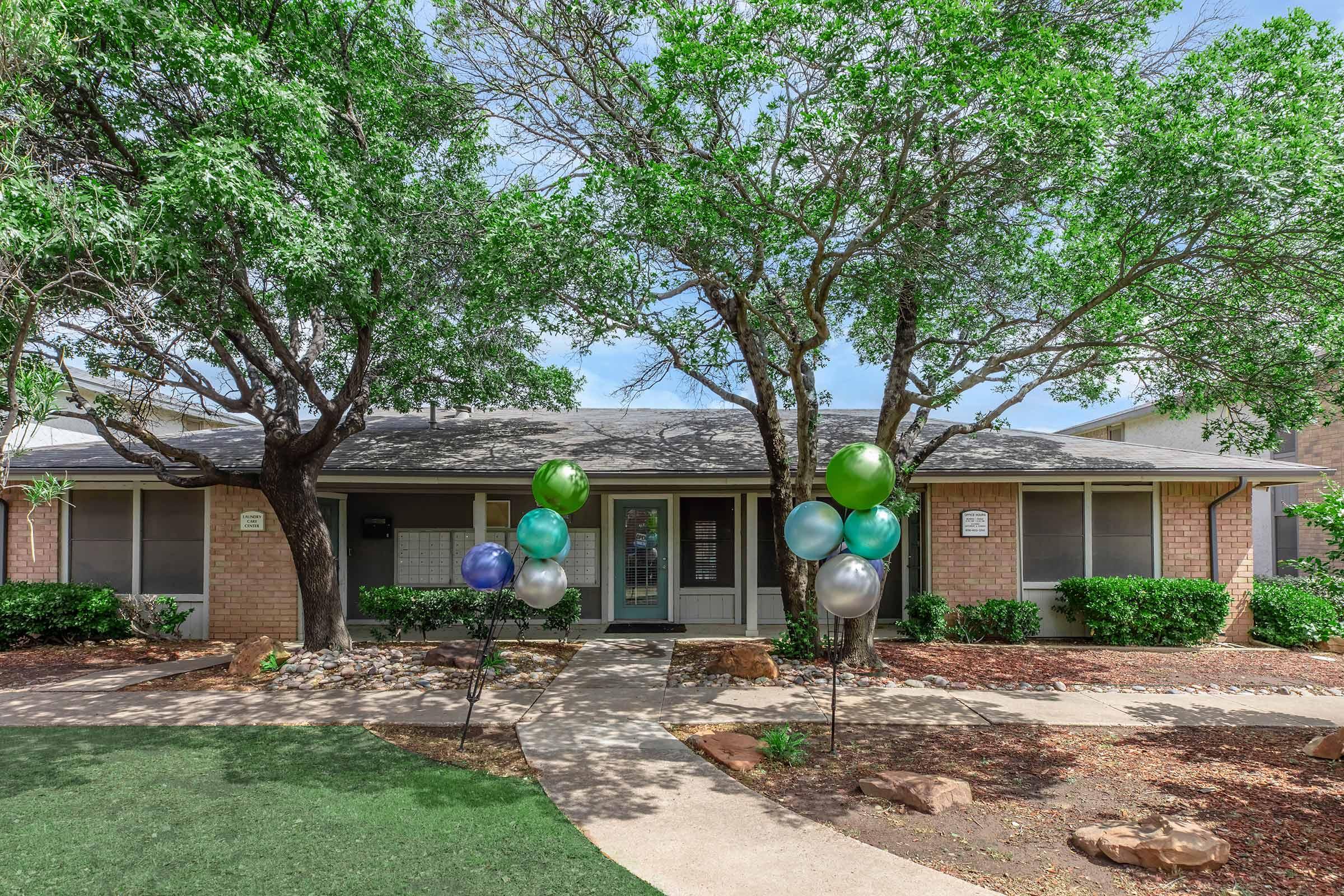 A residential building surrounded by trees and greenery. There are colorful balloons in shades of blue and green at the entrance, and a well-maintained pathway leading to the front door. The setting is bright and inviting, with shrubs and decorative stone features in the landscape.