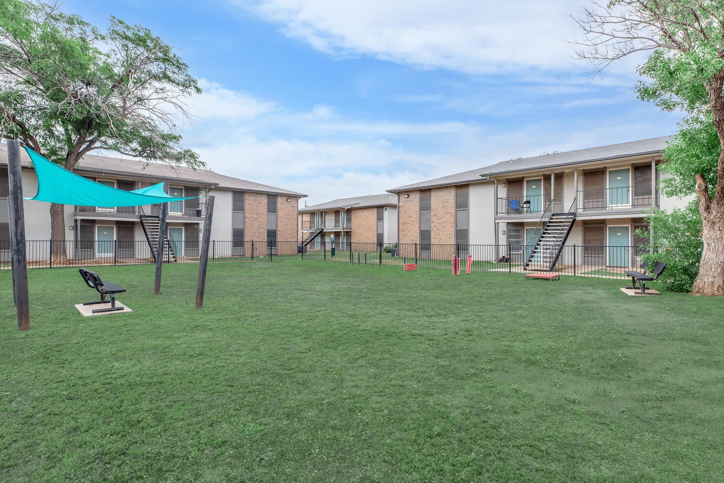 A grassy courtyard with two multi-story apartment buildings on either side. There are playground equipment, including swings and slides, along with black benches and a large tree in the corner. A blue shade structure provides cover in the open space. The sky is clear and bright.