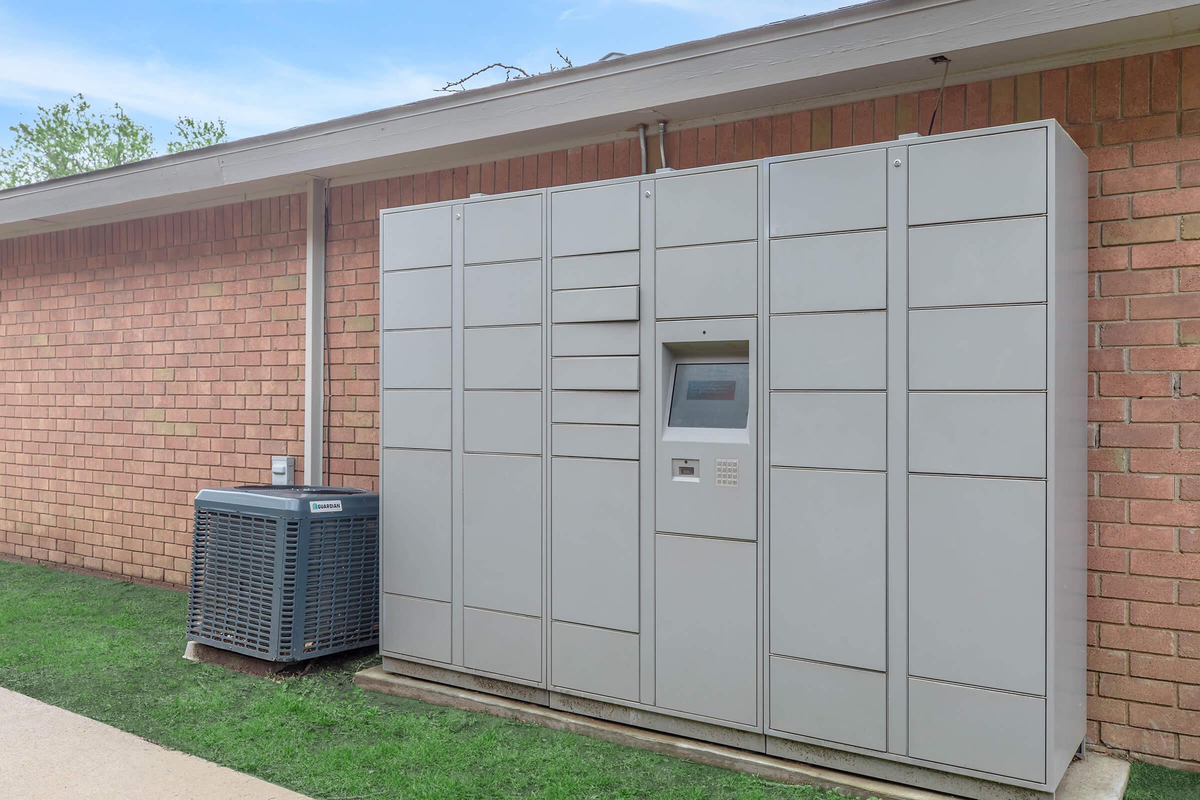 A large, gray outdoor parcel locker system mounted against a brick wall, featuring various compartments for package delivery. An electronic keypad and screen are visible on one side for user interaction. Adjacent to the lockers, there is an air conditioning unit on a grassy area.