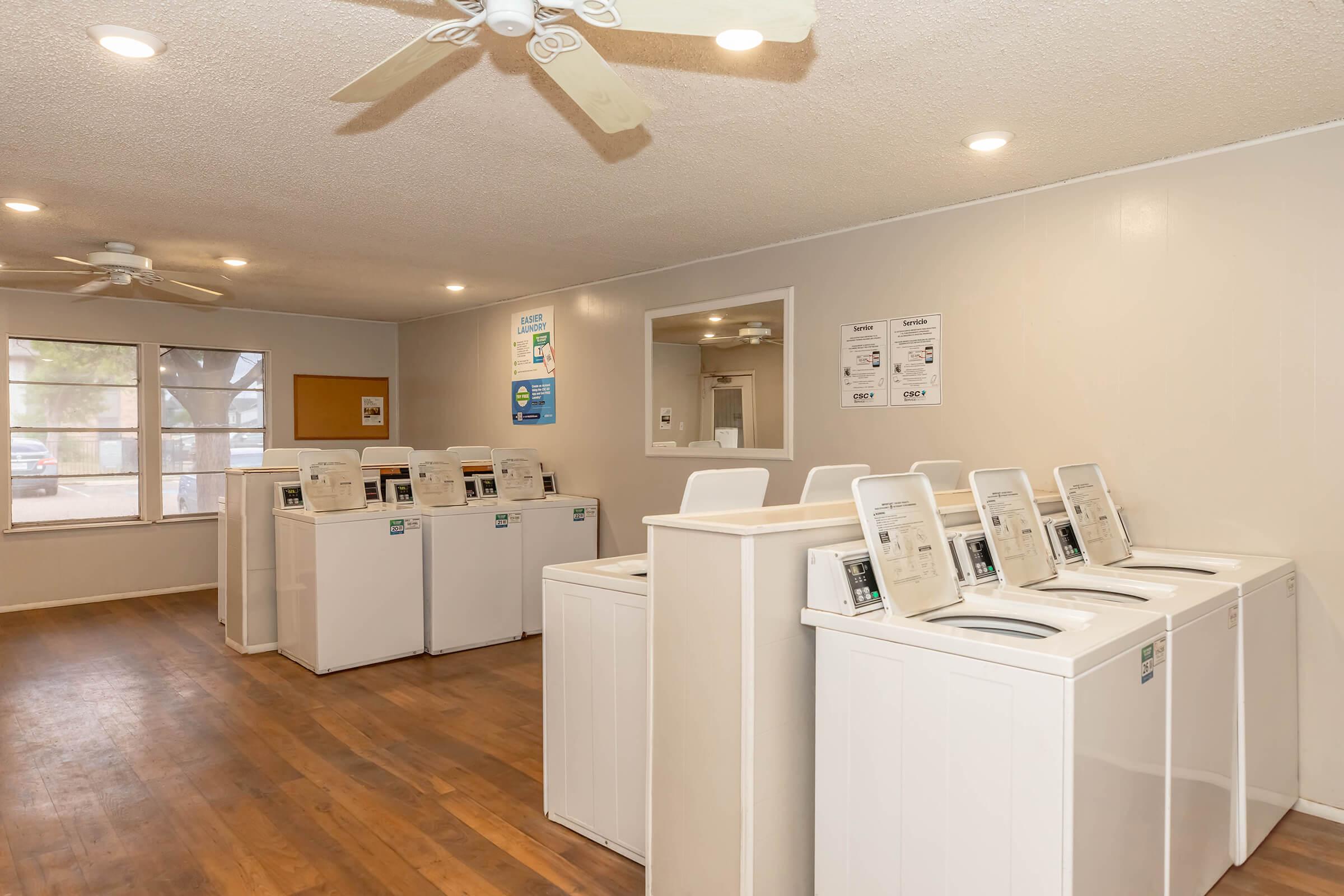 A bright laundry room featuring several white washing machines arranged in rows, with a ceiling fan overhead and natural light coming through large windows. The walls are painted light colors, and there are informational posters on the wall, creating a clean and inviting atmosphere.
