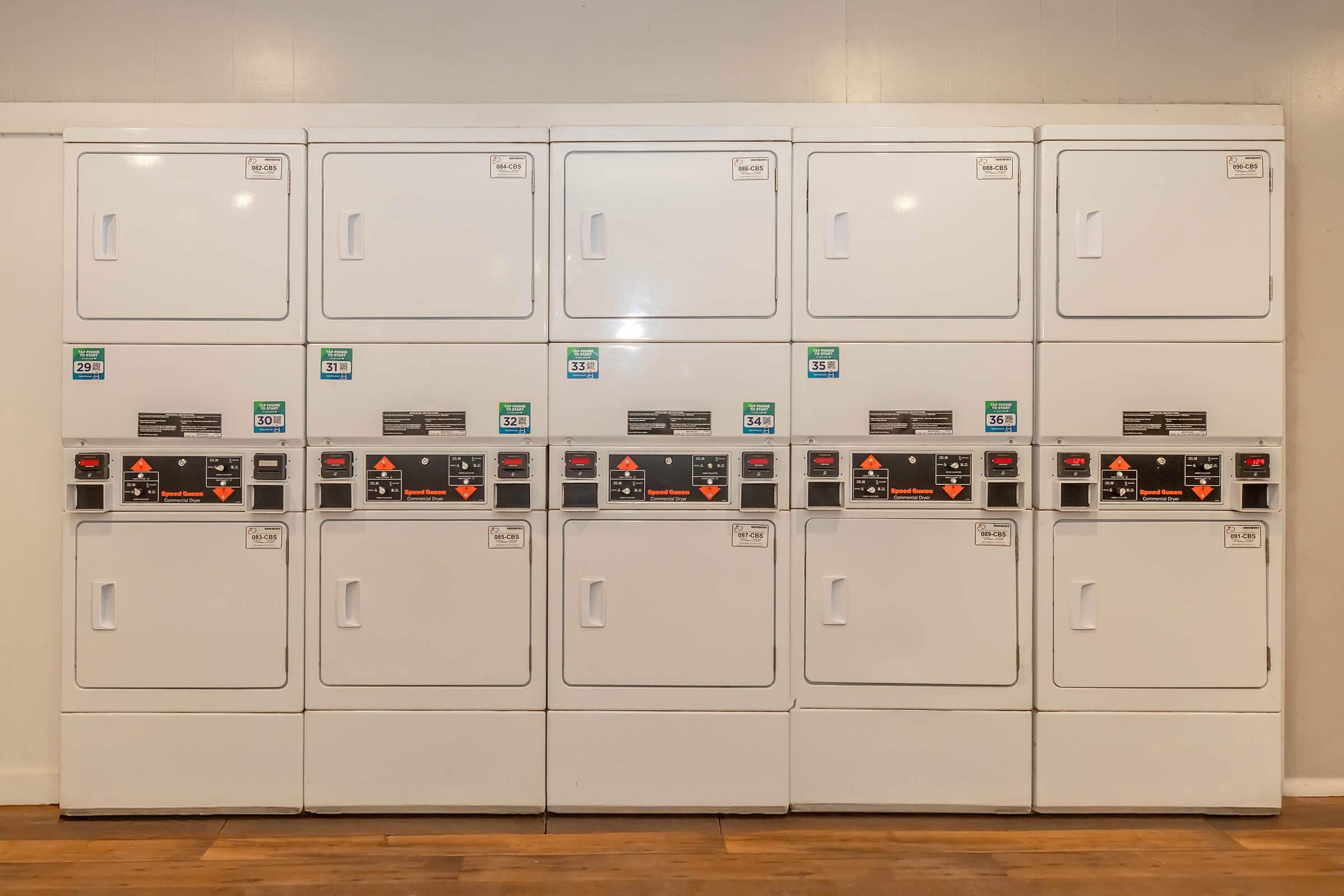 A row of white commercial dryers with multiple settings and coin slots, arranged neatly against a wall. Each dryer features a control panel with labels and indicators for different drying cycles. The setting is clean and well-organized, suitable for a laundromat or shared laundry space.
