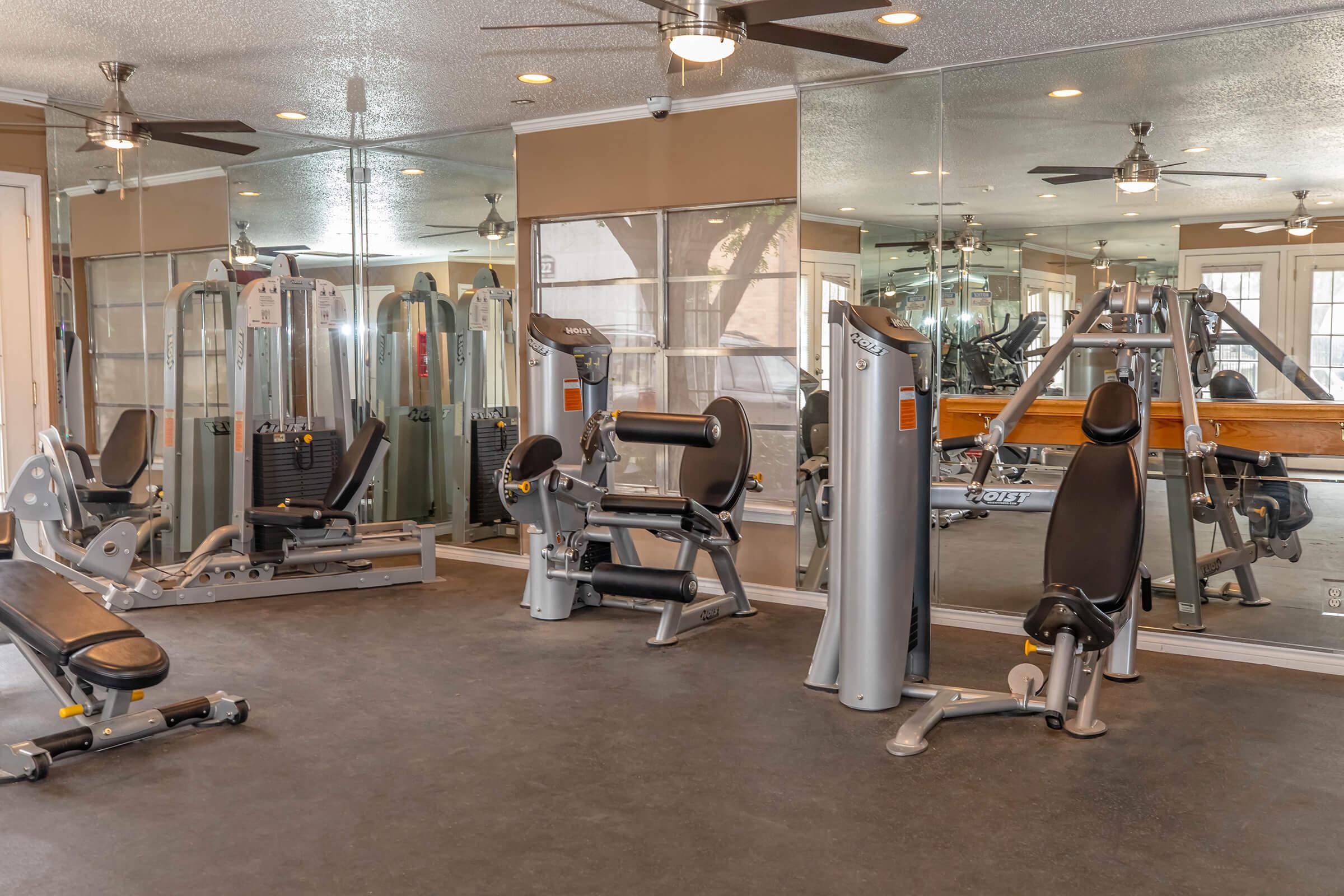 Interior of a gym featuring various exercise equipment, including weight machines and benches. The space is well-lit with mirrors on the walls and ceiling fans. The flooring is dark and the overall design appears modern and inviting, suitable for workouts.