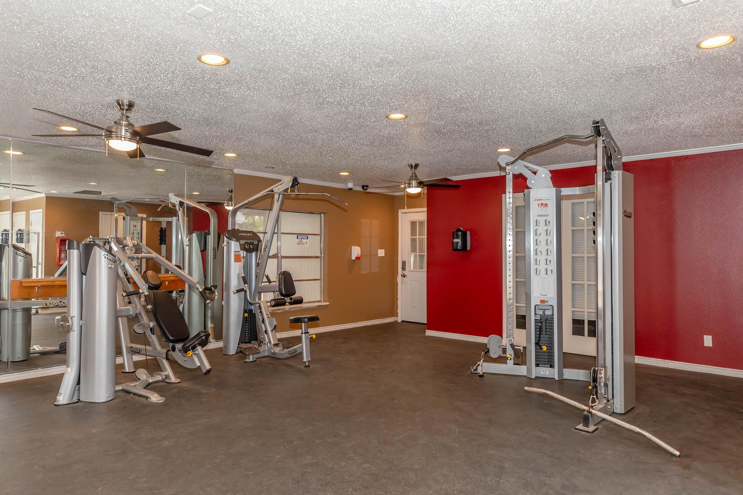A spacious gym with exercise machines, including strength training equipment and cardio stations. The room features a ceiling fan, bright lighting, and mirrors on one wall, creating an inviting space for workouts. A vibrant red accent wall enhances the overall aesthetic.
