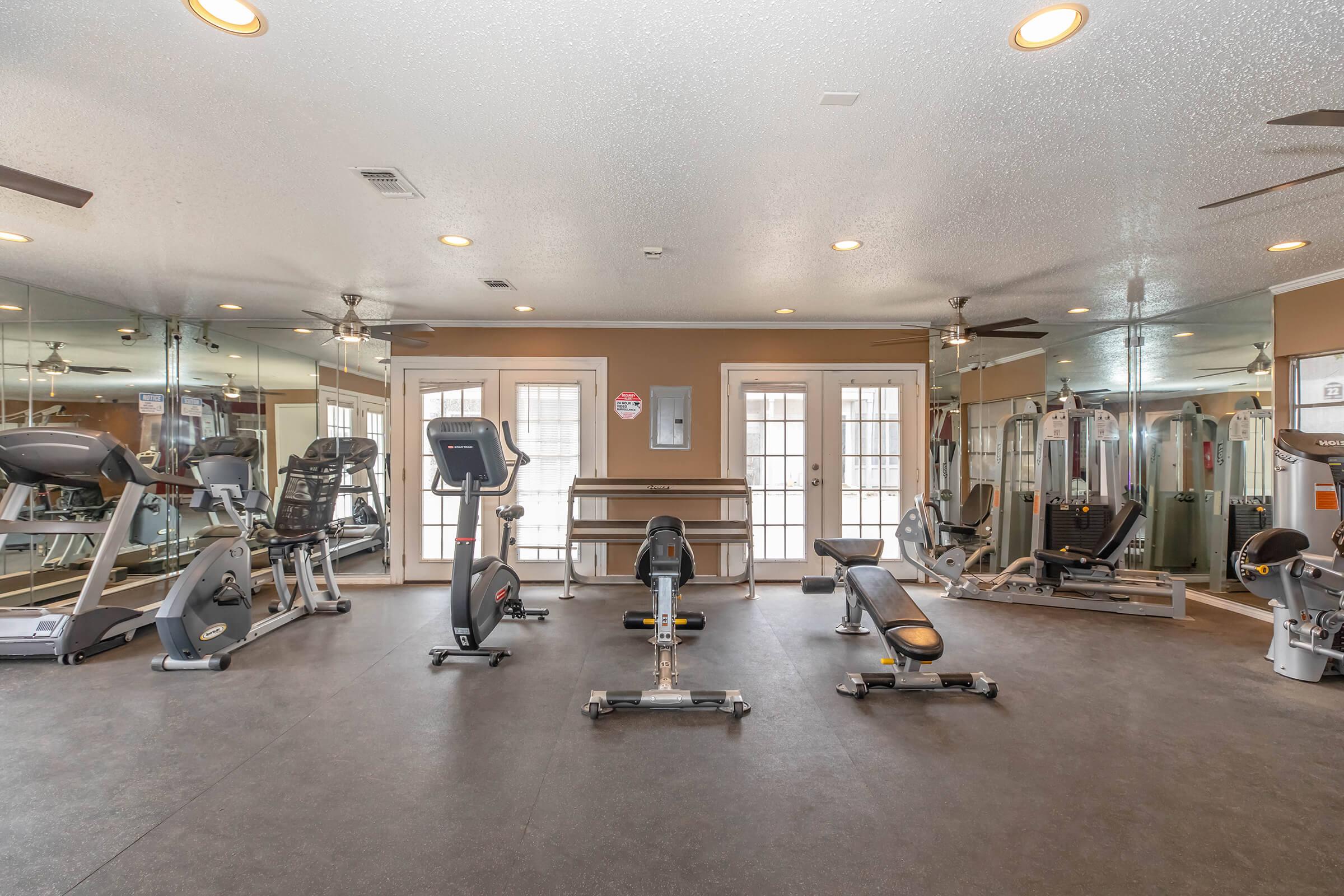 A well-equipped fitness room featuring various exercise machines, including treadmills, stationary bikes, and weightlifting equipment. The space is brightly lit with mirrors on the walls and has a clean, modern design. Double doors lead to an outdoor area, enhancing the workout environment.