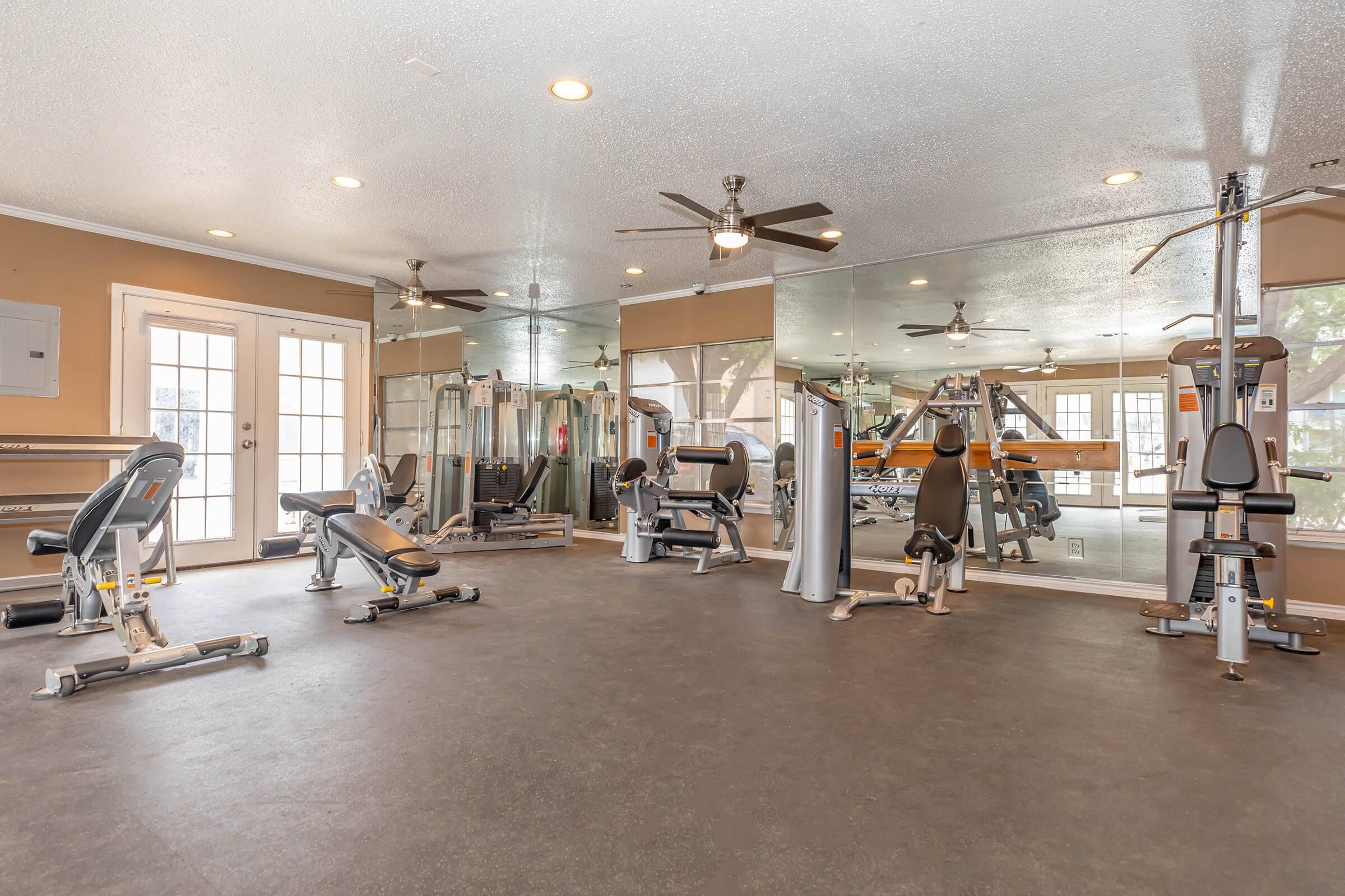 A well-lit, modern gym featuring various exercise equipment, including weight machines, benches, and cardio machines. The space has large mirrors on one wall, ceiling fans, and double glass doors leading outside, creating an inviting atmosphere for workouts.