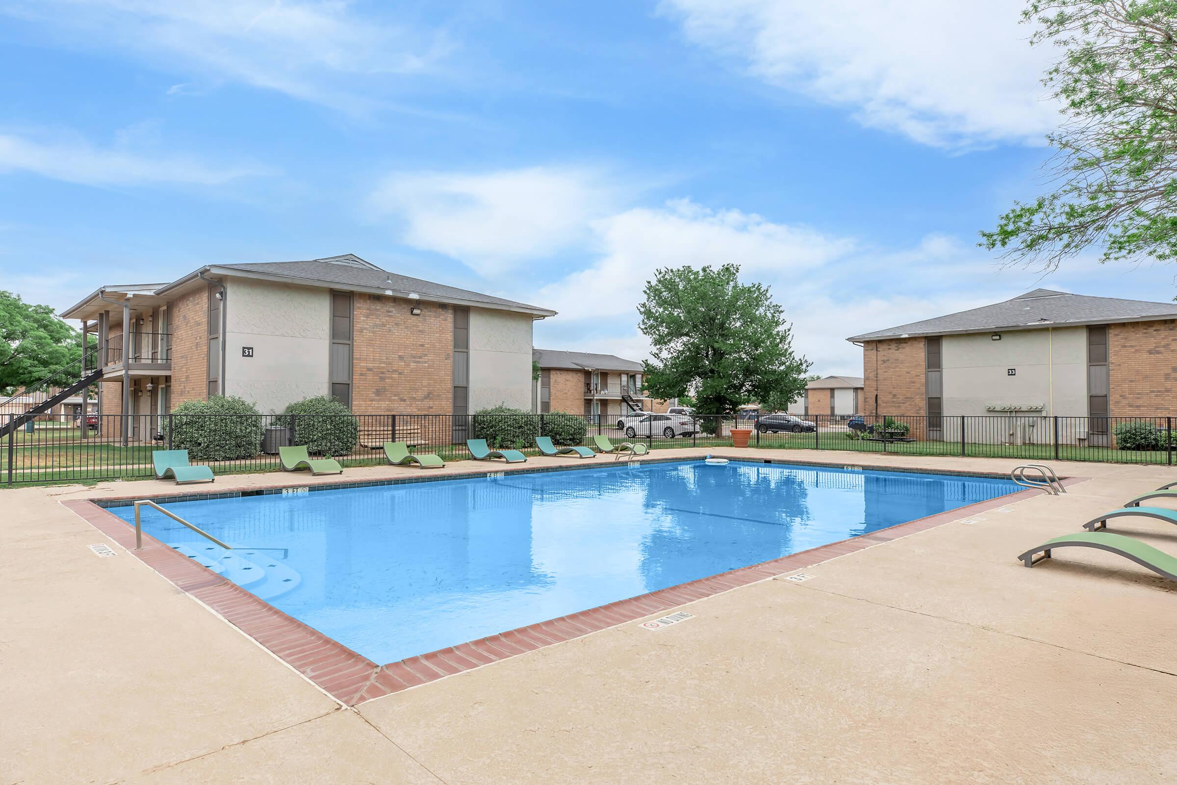 A clear swimming pool surrounded by lounge chairs, with two apartment buildings in the background. The pool area is fenced, and the sky is partly cloudy. Lush green trees and bushes add to the landscaping around the pool area.