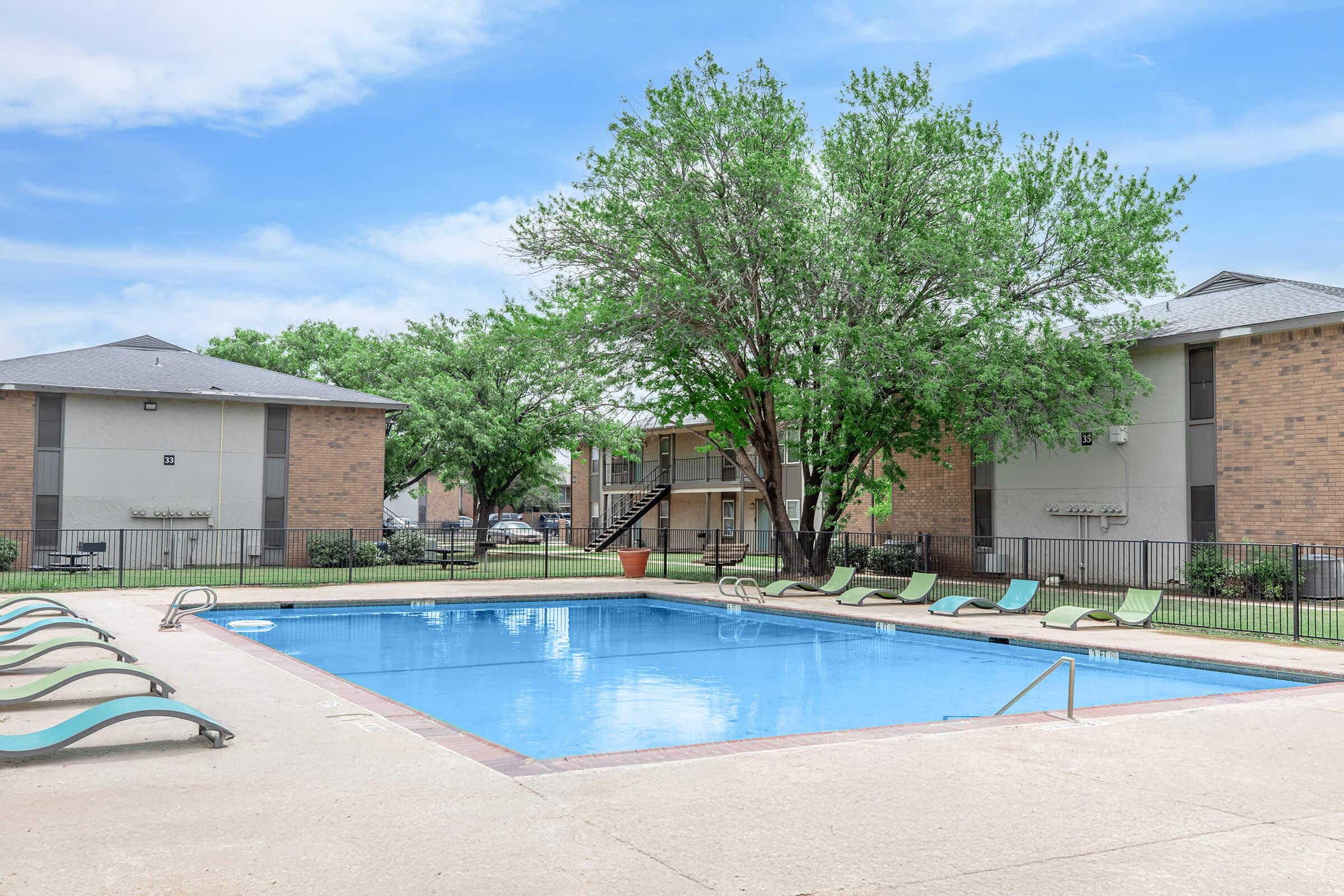 A clear swimming pool surrounded by lounge chairs and green trees, with two multi-story apartment buildings in the background. The sky is blue with a few clouds, creating a bright and inviting atmosphere.