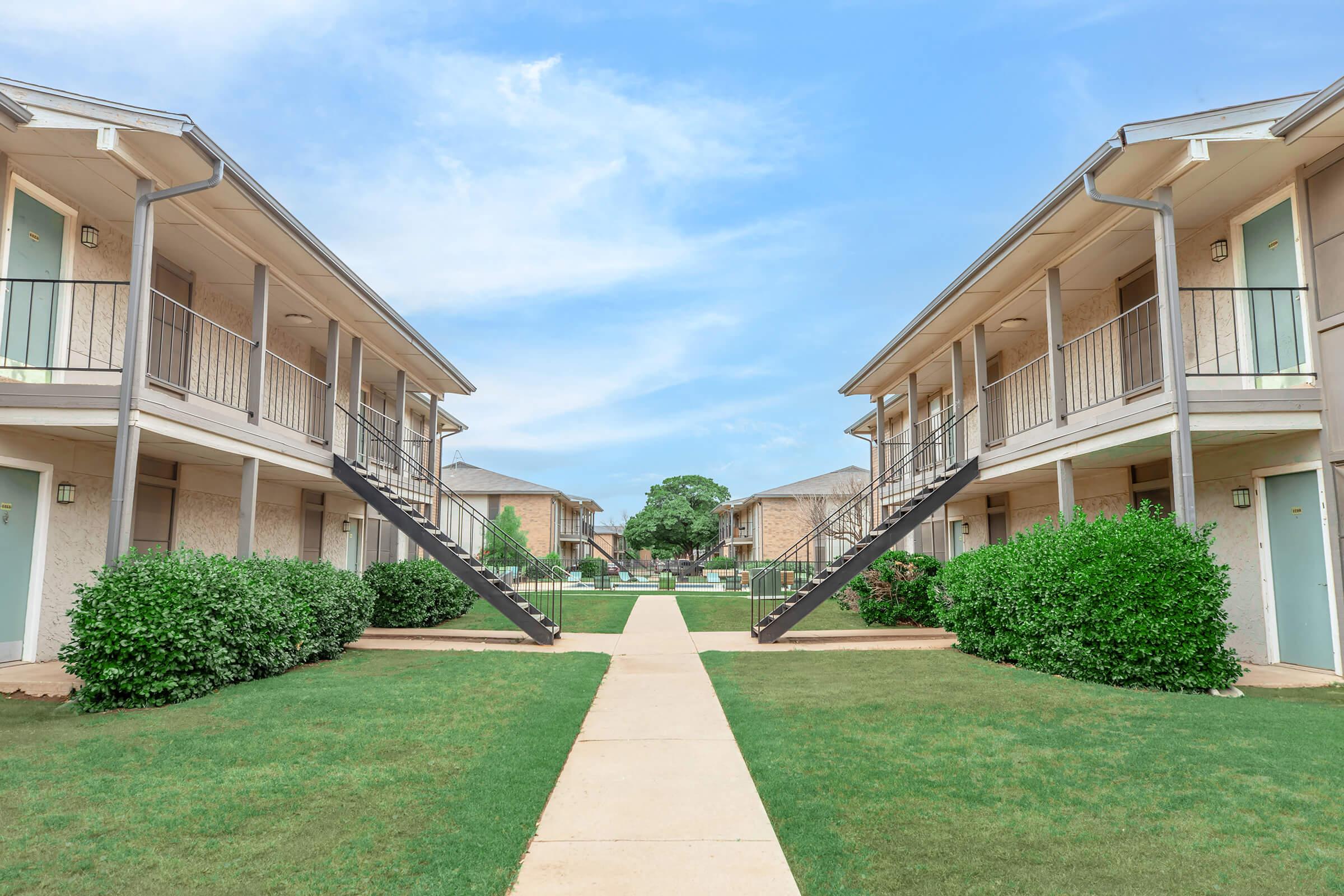 Two parallel buildings with balconies and stairs leading up to them, separated by a neat pathway. The lawn is well-maintained with green shrubs on either side. The sky is clear with a few clouds, creating a serene atmosphere in a residential area.