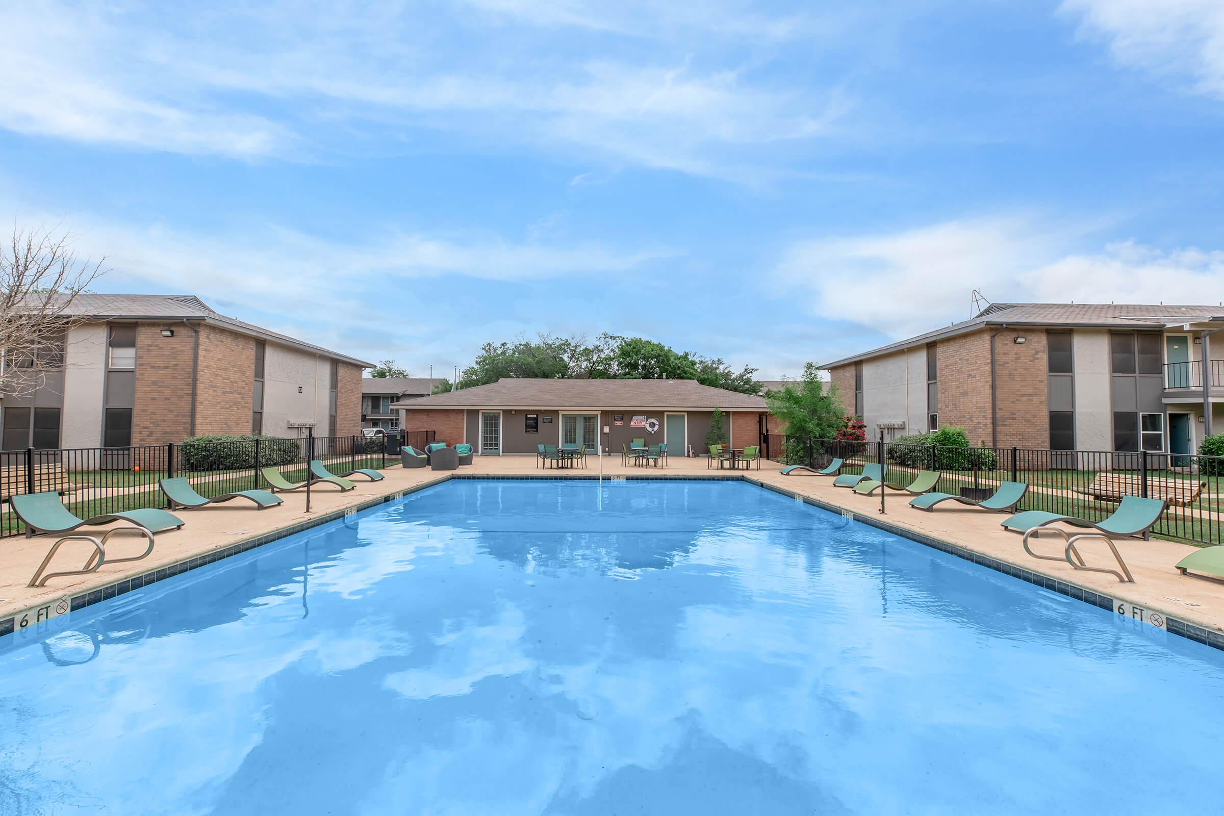 A clear, blue swimming pool surrounded by a well-maintained area. Lounge chairs are positioned along the pool's edge, with a clubhouse visible in the background. The scene is framed by two brick buildings and features a bright, sunny sky.