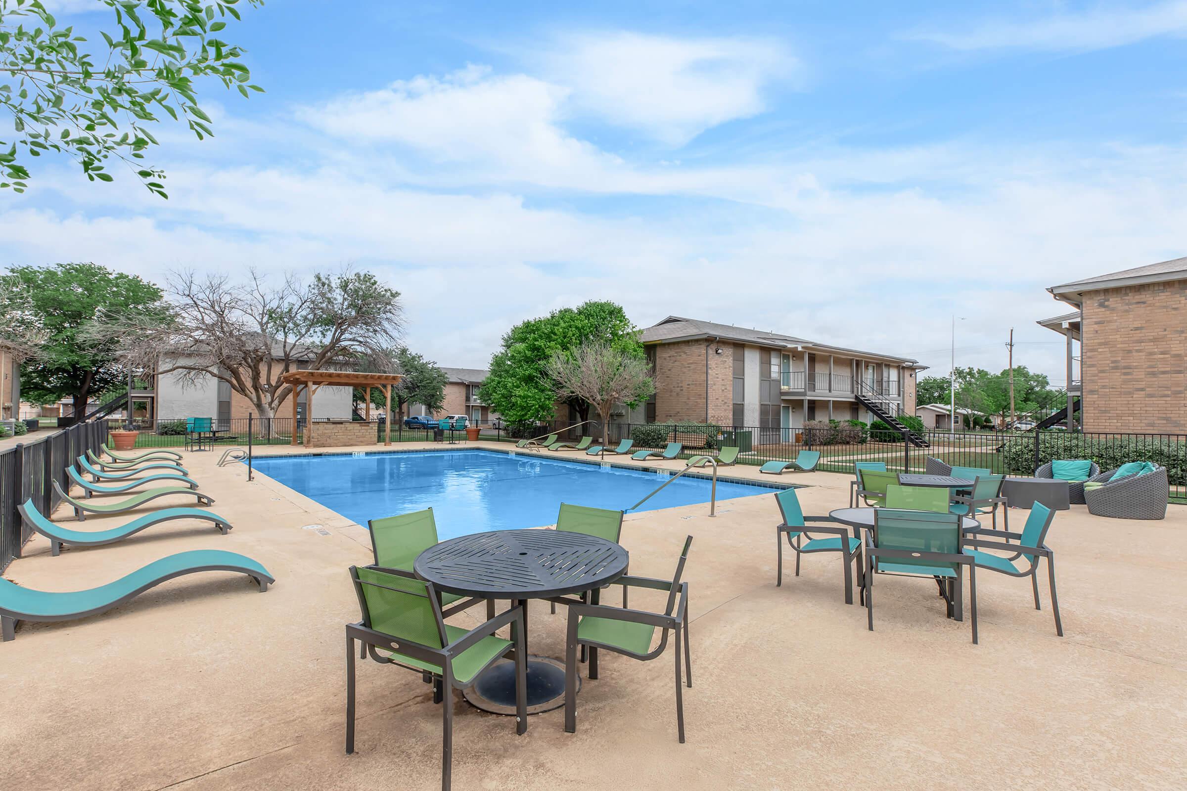 A clear blue swimming pool surrounded by lounge chairs and tables with green chairs. In the background, there are buildings and trees under a partly cloudy sky. The area is well maintained, inviting relaxation and leisure.