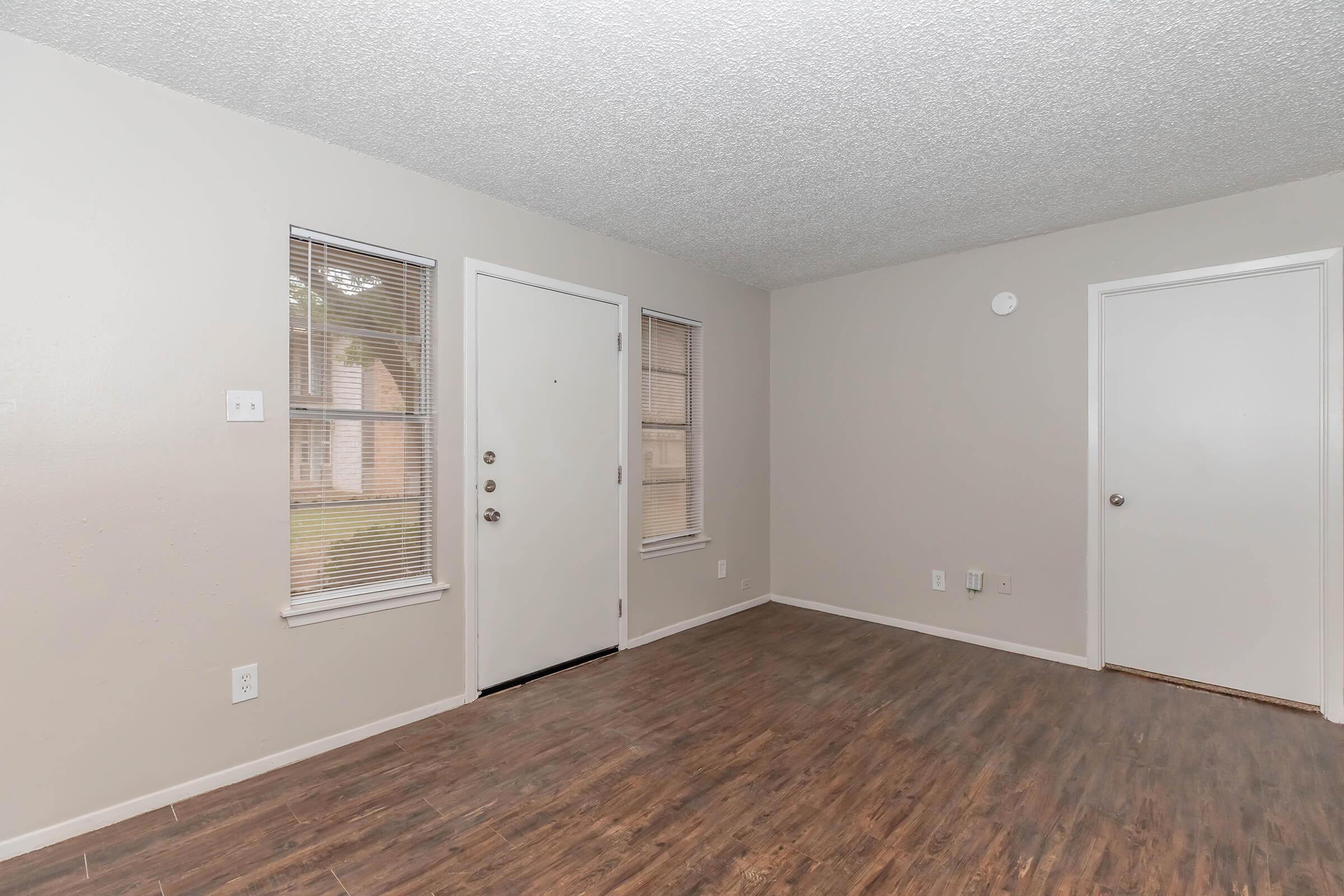 A well-lit living room featuring a textured ceiling, wooden flooring, and neutral-colored walls. It includes a front door with multiple locks, two windows with blinds, and a door leading to another room. The space is empty, offering a clean and inviting atmosphere.