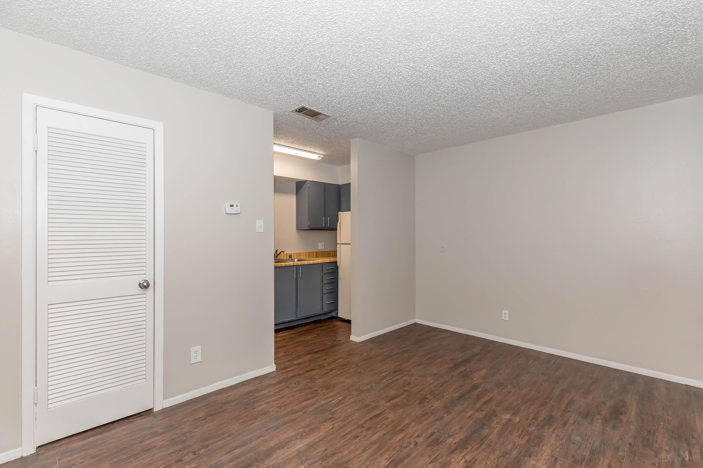 A well-lit living space with a textured ceiling, featuring a neutral-colored wall. There is a white door on the left leading to another room, and an open kitchen area in the background with gray cabinets and a countertop. The floor is a wood-like laminate, enhancing the modern feel of the room.