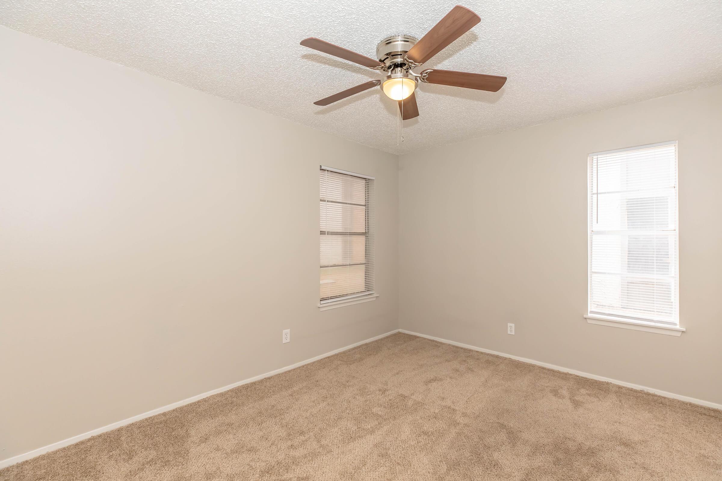 Empty room with beige walls and light brown carpet. Two windows with blinds allow natural light. A ceiling fan with wooden blades is installed for ventilation. The space appears to be well-maintained and ready for furnishings.