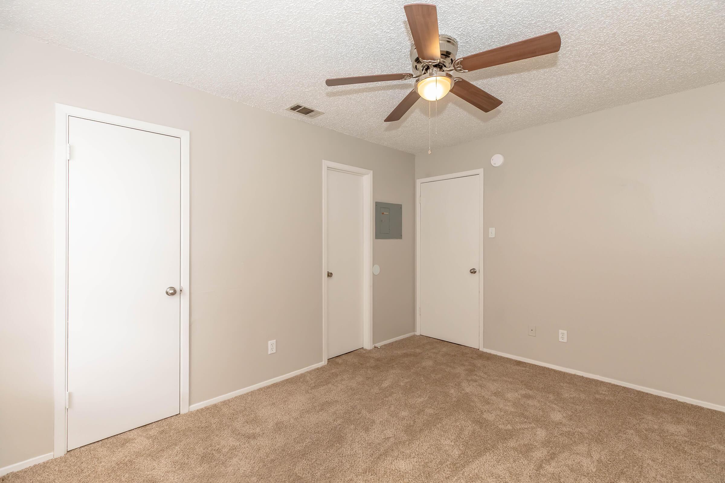 A vacant room with light beige carpet and textured ceiling. Two white doors are visible on the left and a third door is on the right. A ceiling fan is mounted in the center, providing light. The walls are painted in a neutral gray color. The space appears well-lit and tidy.