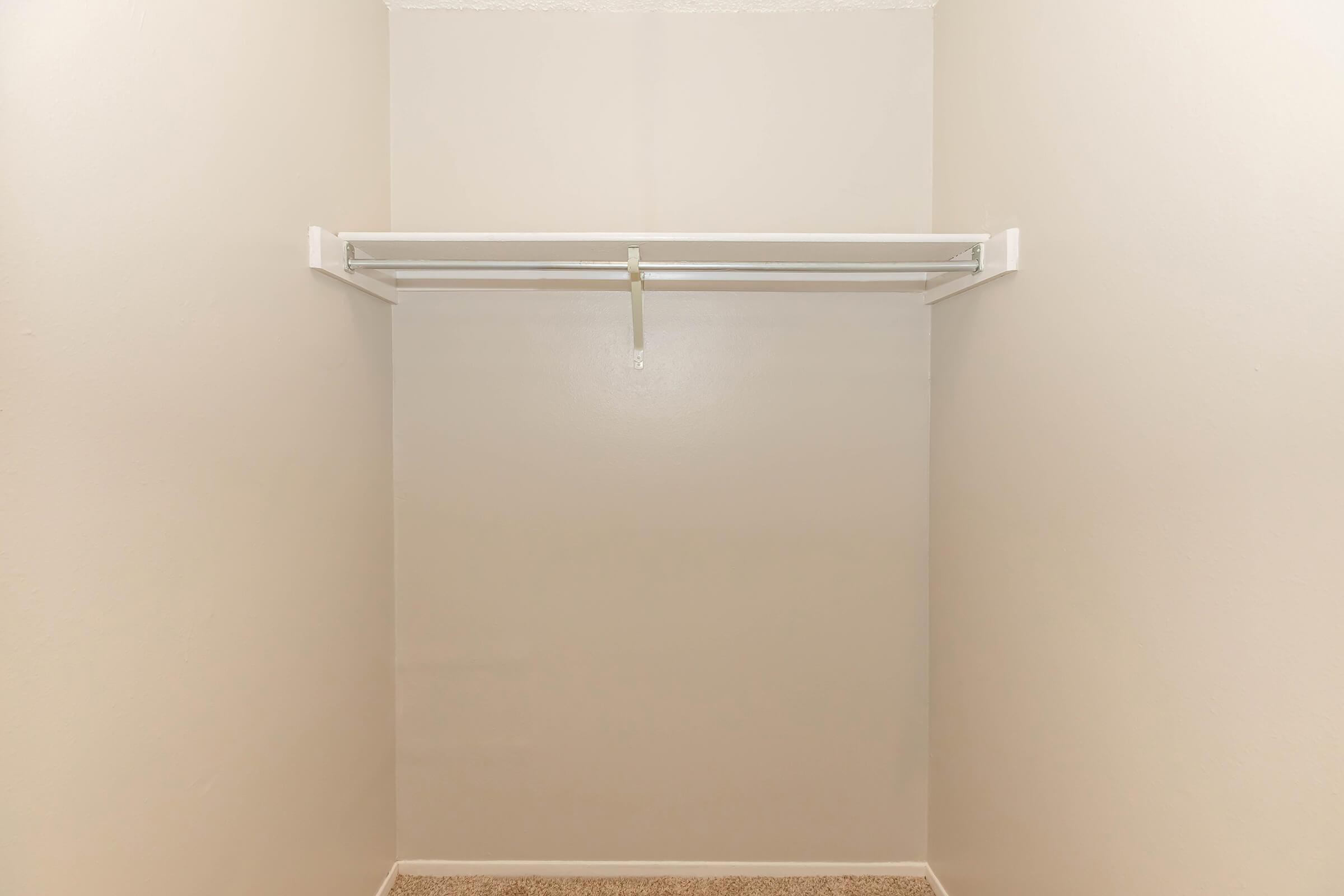 Empty closet with beige walls and a white hanging rod. The floor is covered in light-colored carpet, and there are no items or shelves visible inside. The overall appearance is clean and minimalistic.
